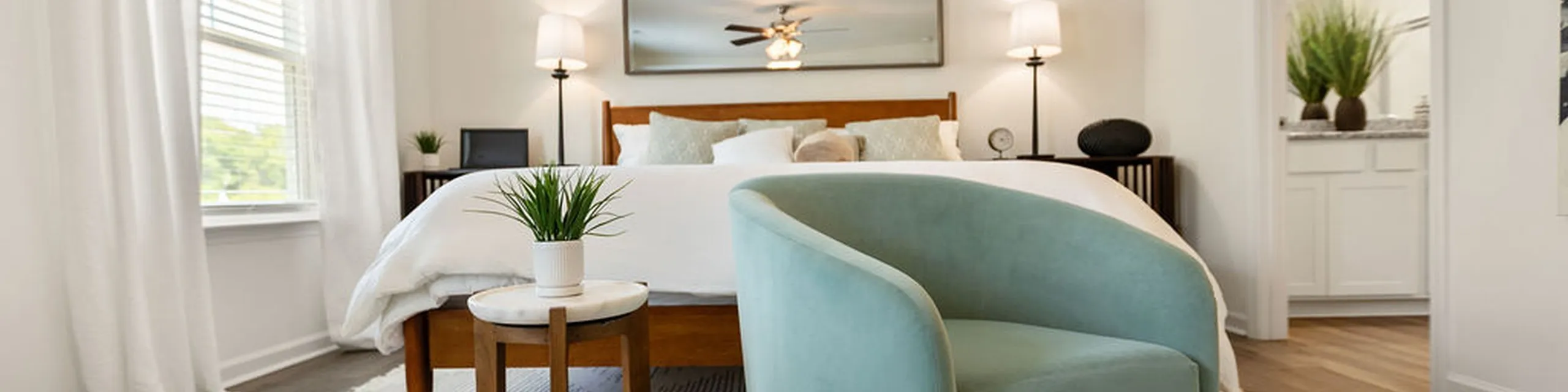Elegant bedroom featuring a cozy bed, stylish teal chair, and bright natural light from large windows, offering a serene atmosphere.