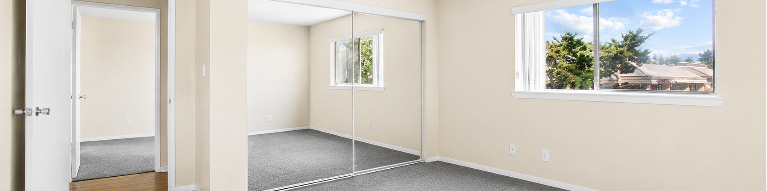 Bright and airy bedroom with large mirrored closet doors, plush gray carpeting, and a sunlit window offering scenic views.