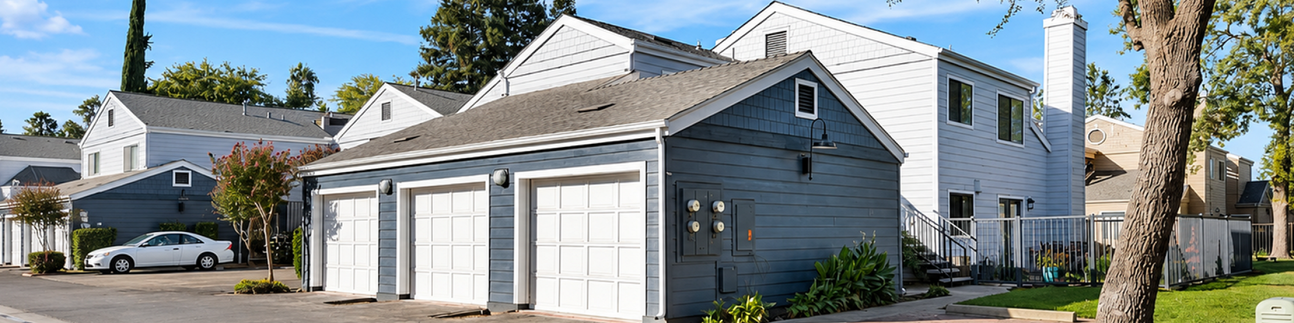 Charming suburban complex featuring modern townhouses, each with a spacious garage and surrounded by lush greenery on a bright sunny day.