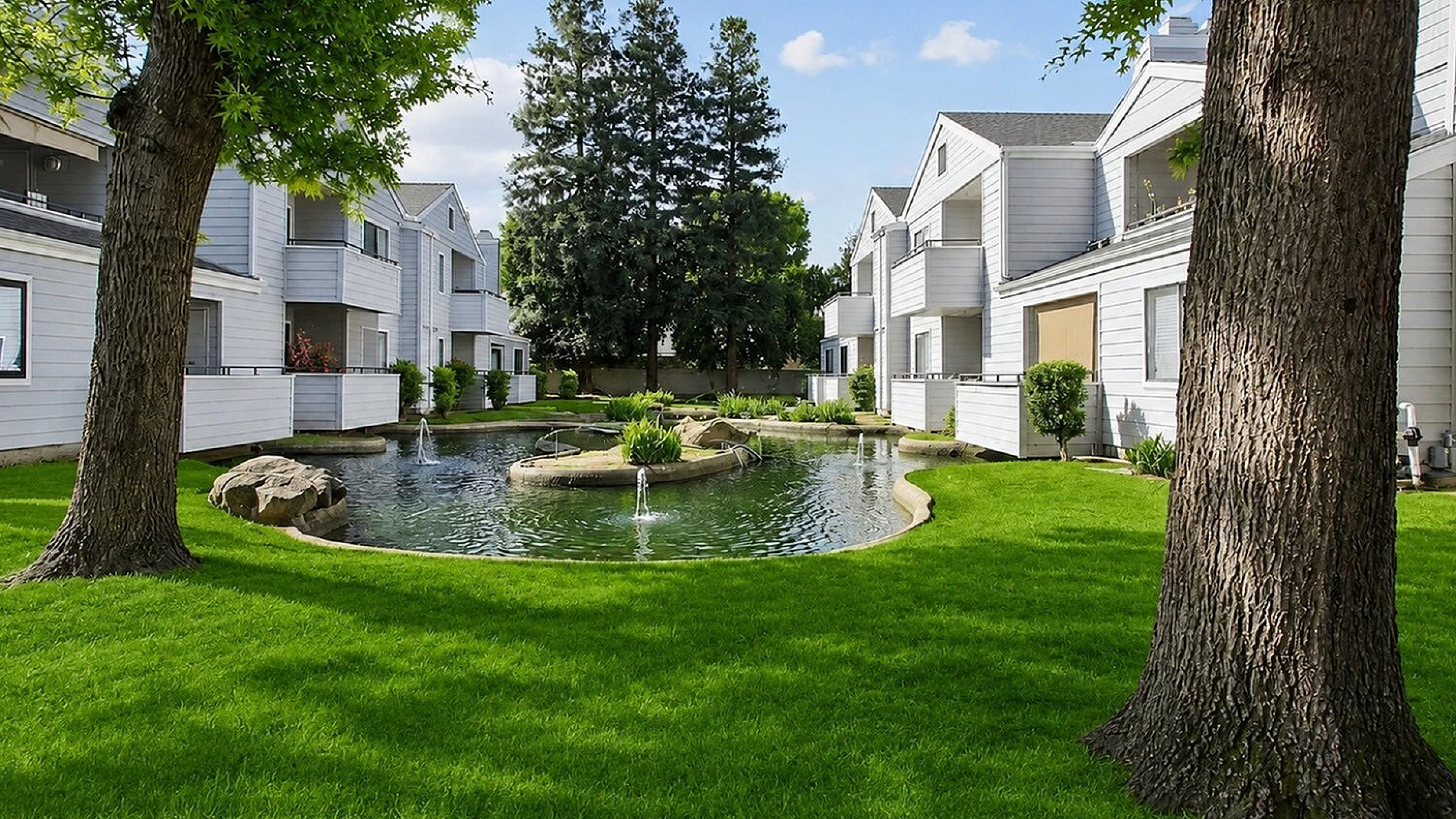 Charming apartment complex with elegant white facades, lush green lawn, and serene pond with fountains.