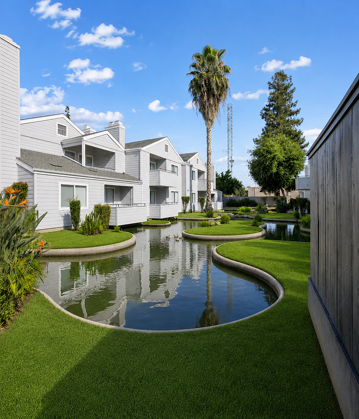 Charming apartment complex with serene landscaped gardens and reflective pond, framed by clear blue skies.