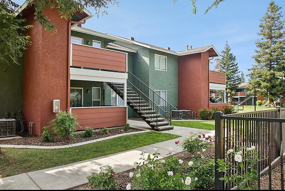 Charming apartment building with a modern exterior, surrounded by lush greenery and vibrant flowers, featuring an inviting balcony and convenient outdoor staircase.