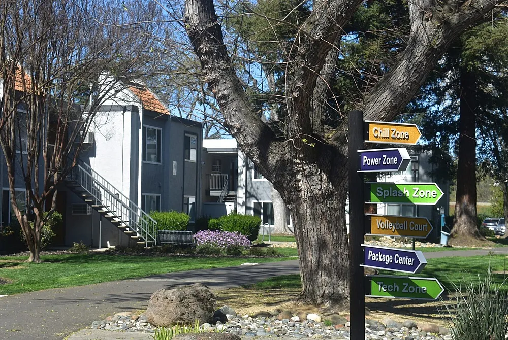 Charming apartment complex with lush greenery and inviting pathways, featuring vibrant directional signs to amenities like Chill Zone and Splash Zone.