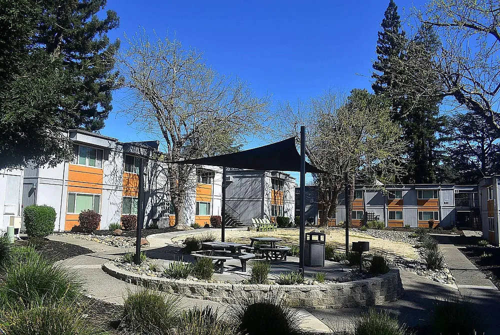 Charming apartment complex exterior showcasing modern architecture, lush landscaping, and inviting picnic area under a sun sail.