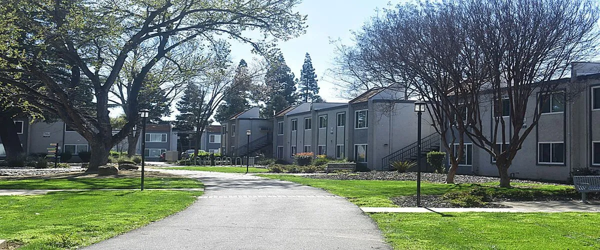 Charming apartment complex nestled in a serene, green landscape with mature trees and a welcoming walkway.