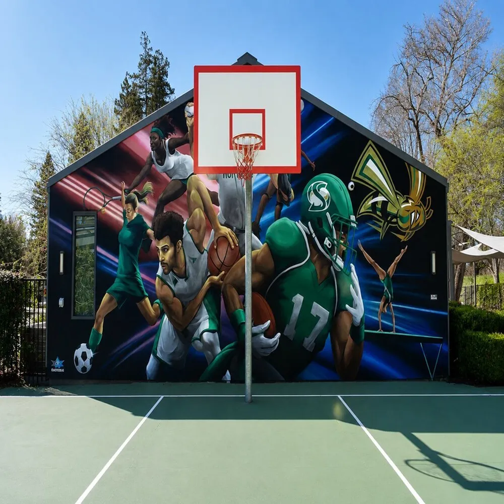 Vibrant sports mural on exterior wall of a building, featuring dynamic athletes from various sports with a basketball hoop in the foreground, creating an energetic community atmosphere.