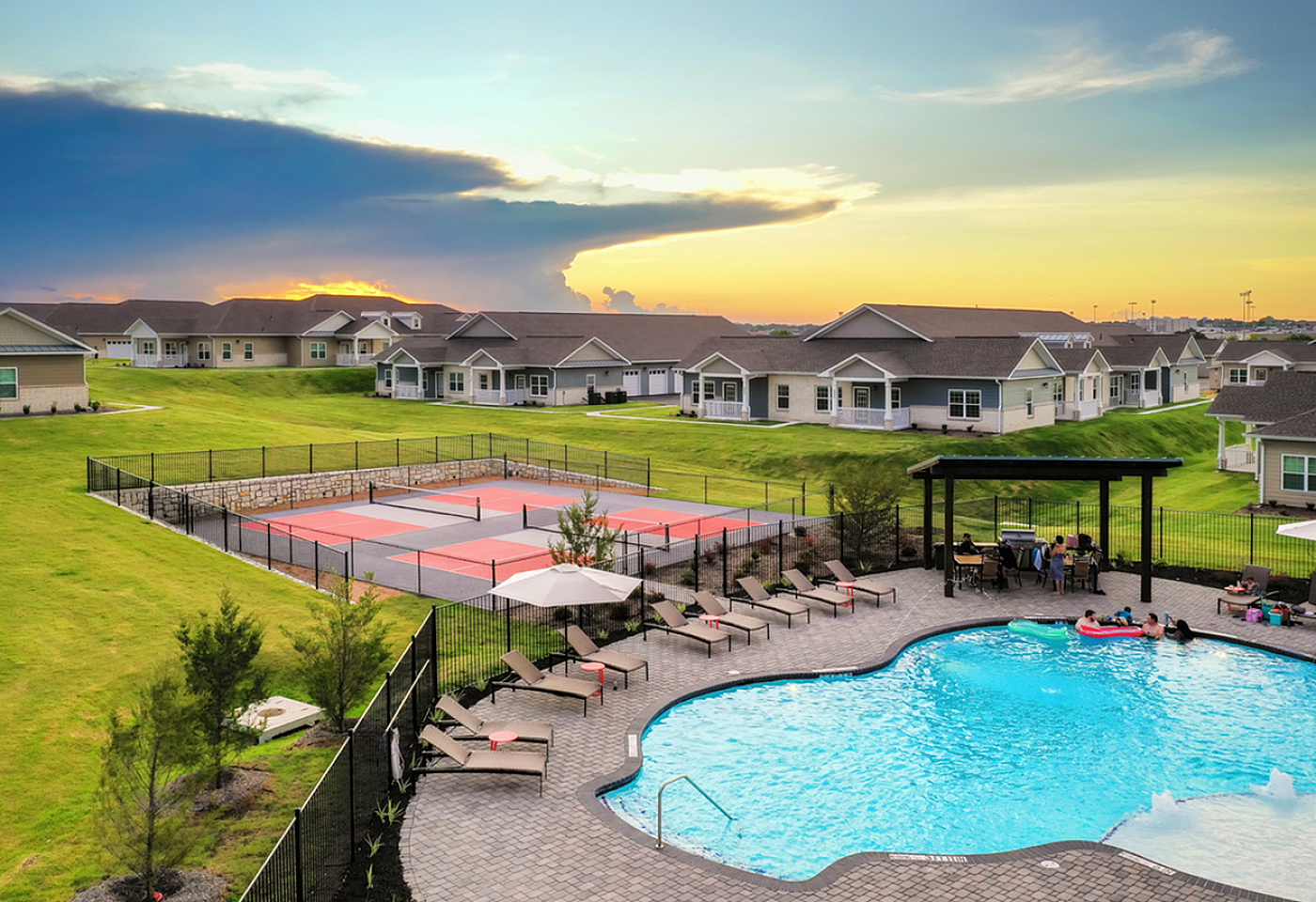 Charming residential community with modern homes, relaxing pool area, and vibrant tennis court set against a stunning sunset backdrop.