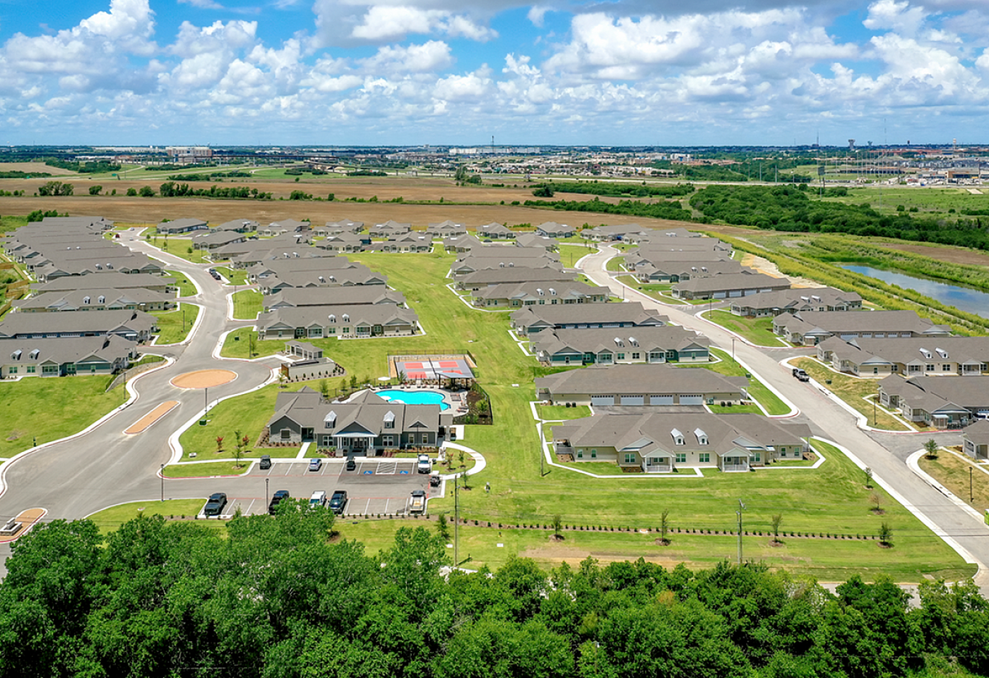 Expansive aerial view of a modern apartment complex with lush green surroundings, tennis court, and sparkling swimming pool under a bright blue sky.