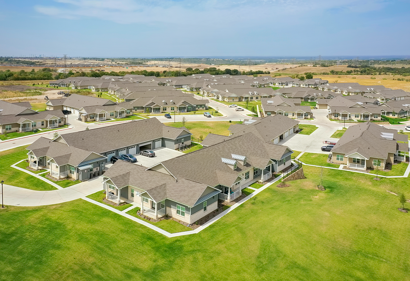 Aerial view of a modern suburban neighborhood featuring charming single-story homes with manicured lawns, spacious driveways, and scenic surroundings under a bright blue sky.