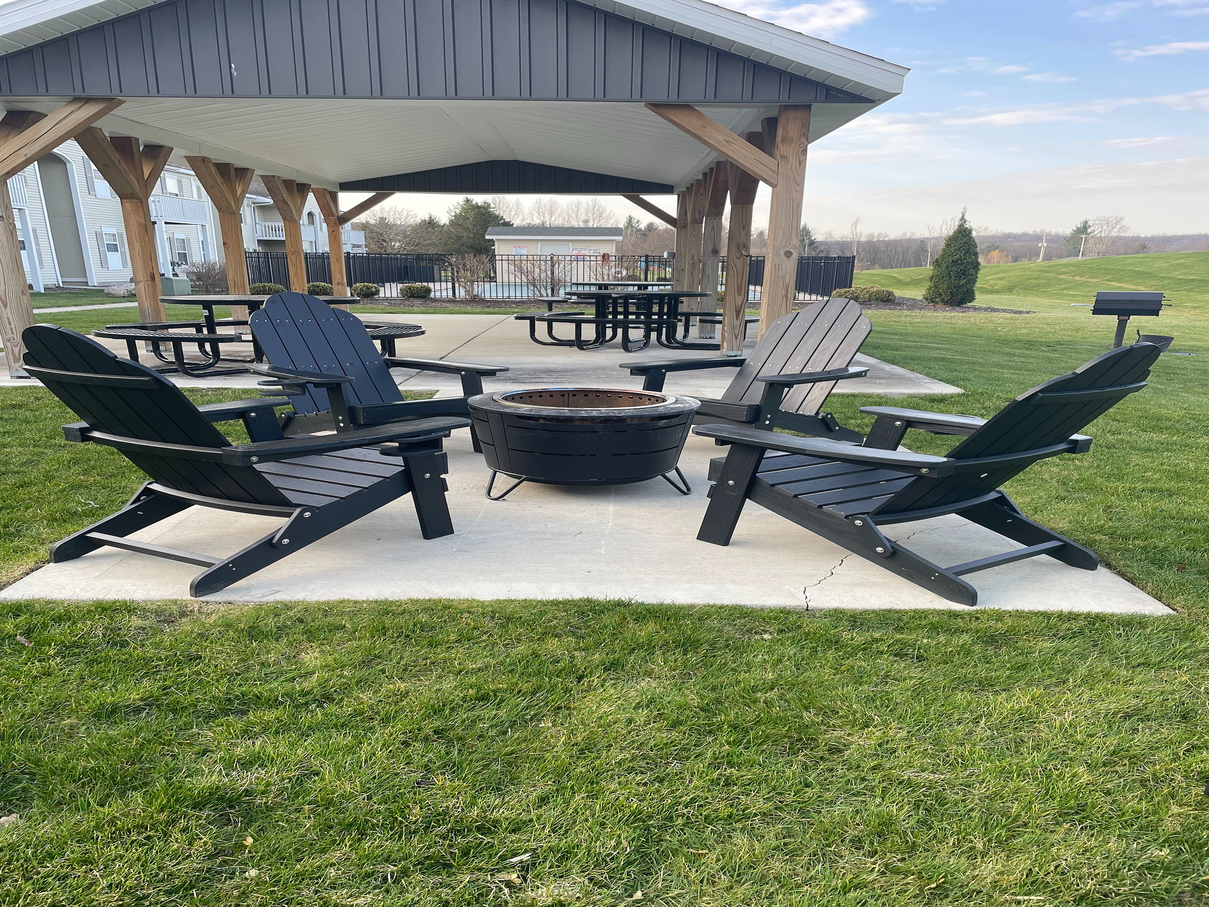 Charming outdoor area with Adirondack chairs around a fire pit under a cozy pavilion, surrounded by lush green grass and picnic tables.