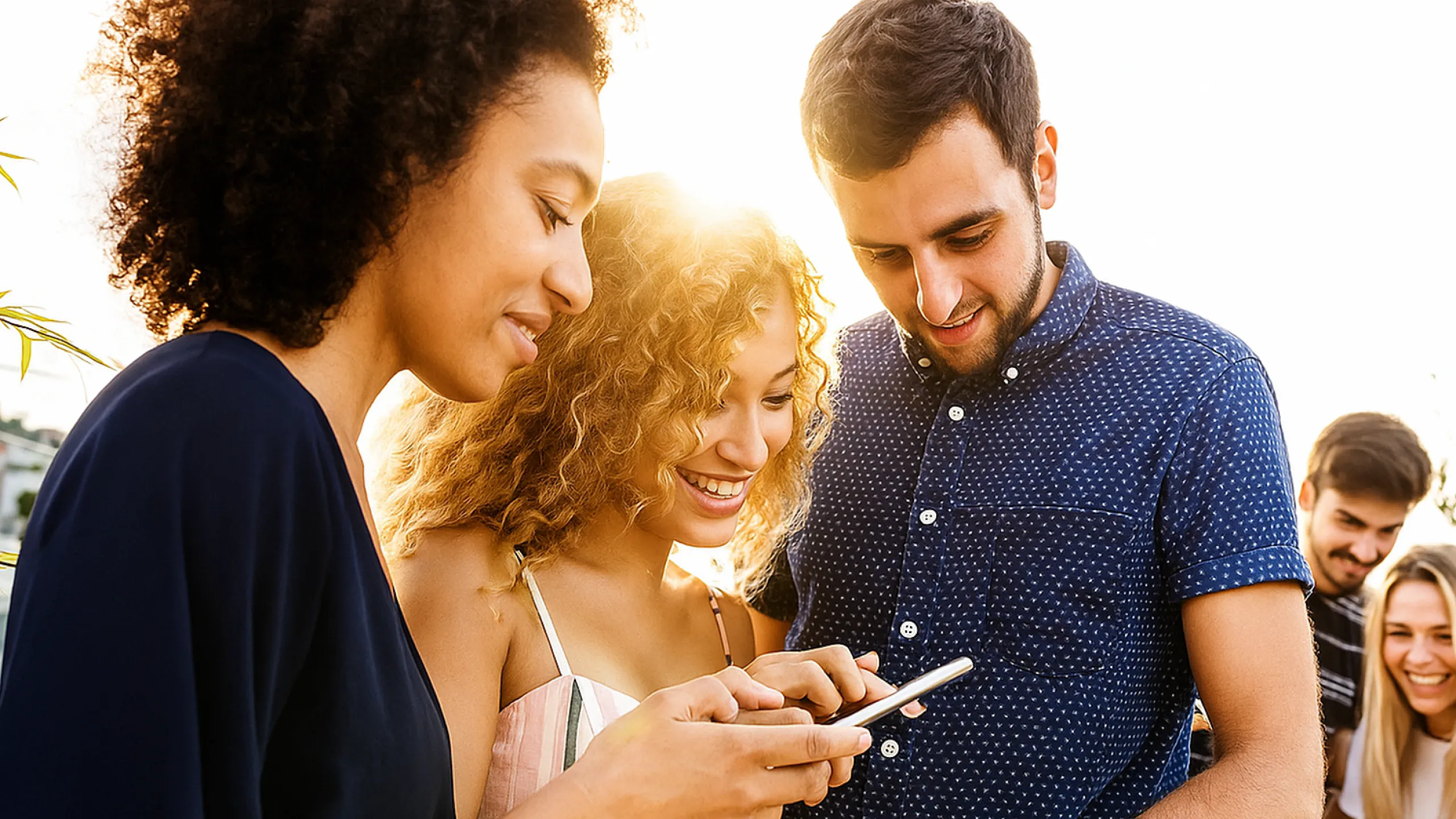 Group of friends enjoying a sunny day outdoors, gathered around a smartphone, sharing joyful moments and laughter.