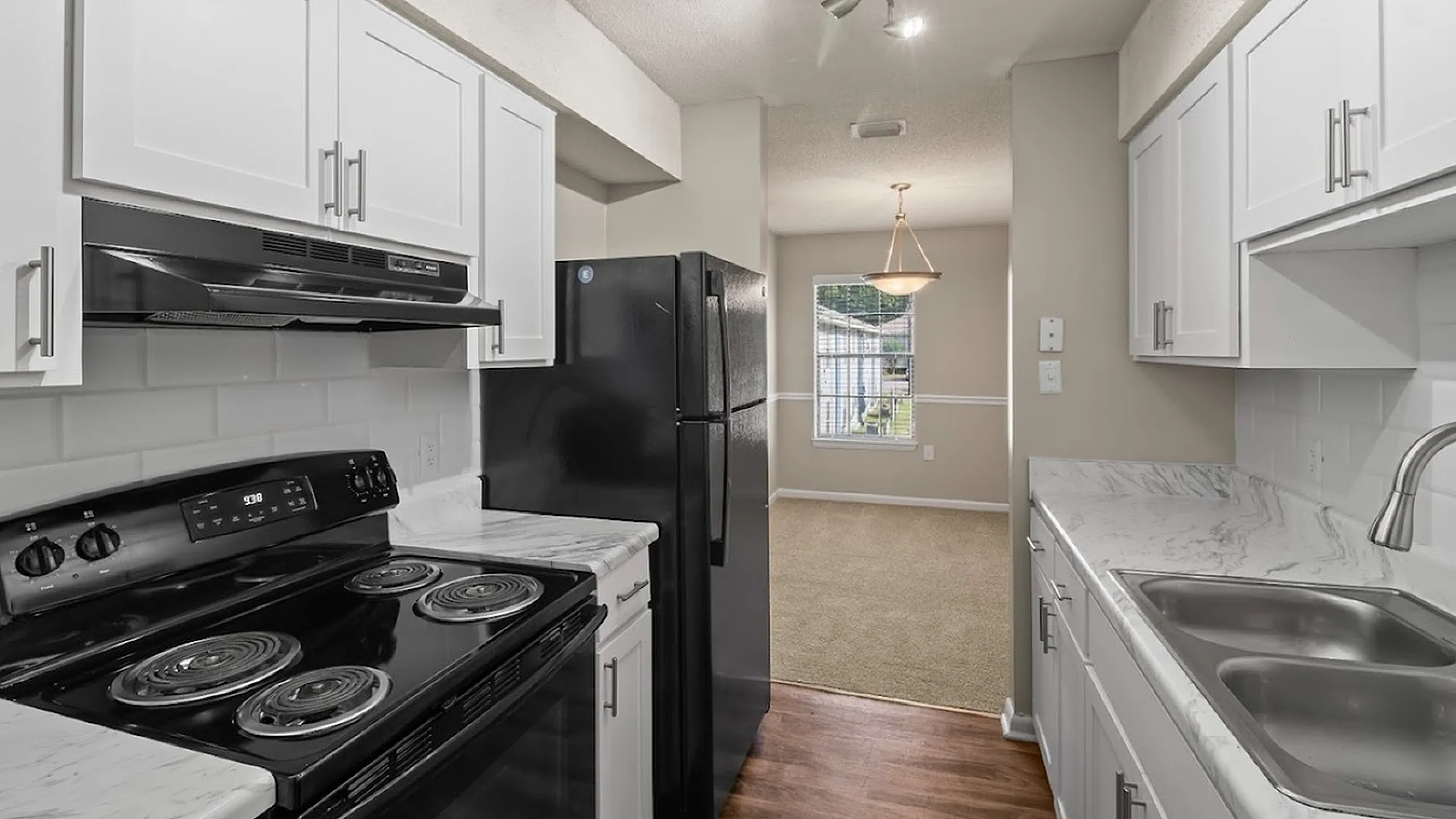 Modern kitchen with sleek white cabinets, black appliances, and hardwood flooring leading to a bright dining area.