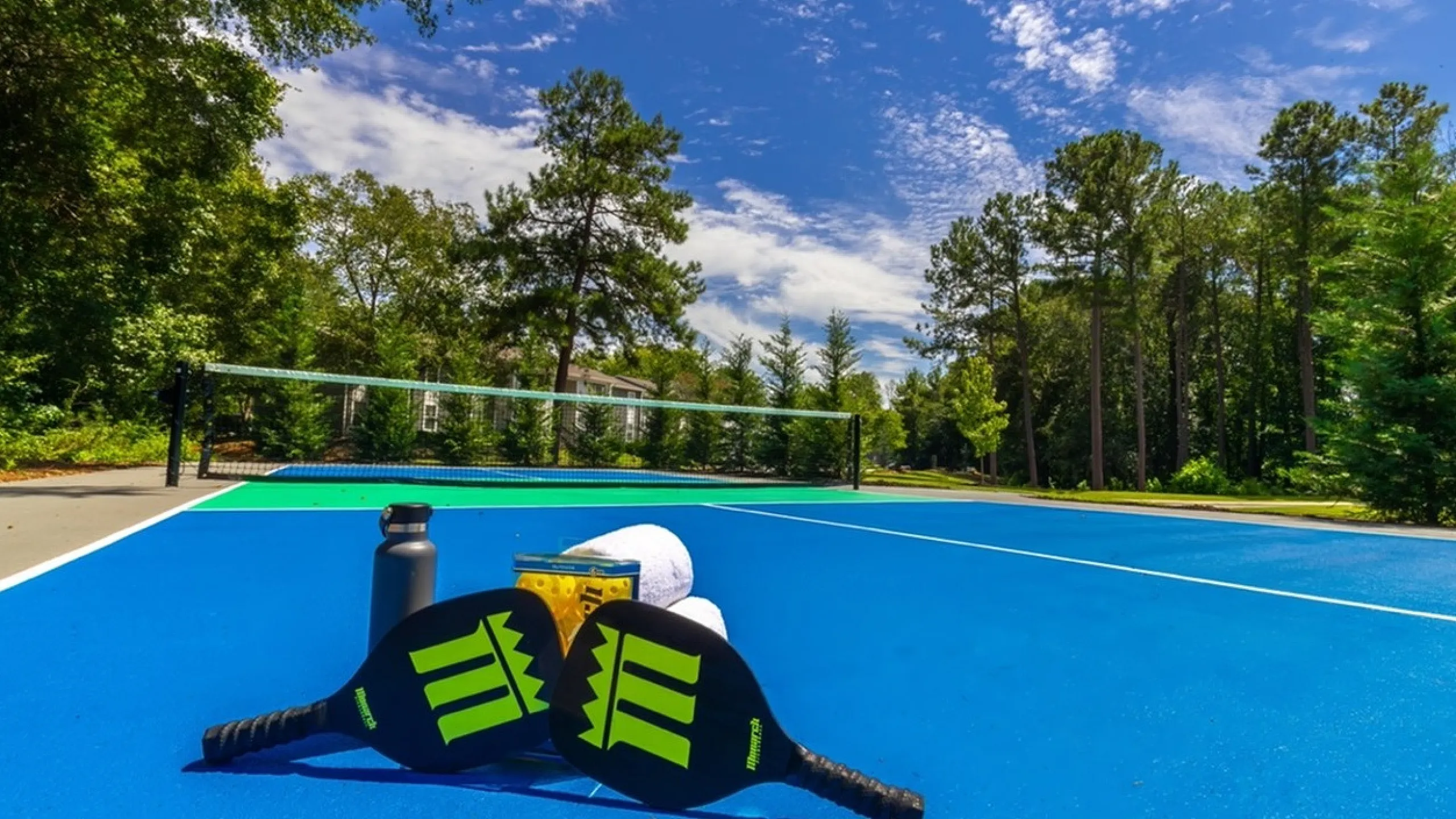 Vibrant tennis court surrounded by lush greenery under a clear blue sky with tennis equipment on the ground.
