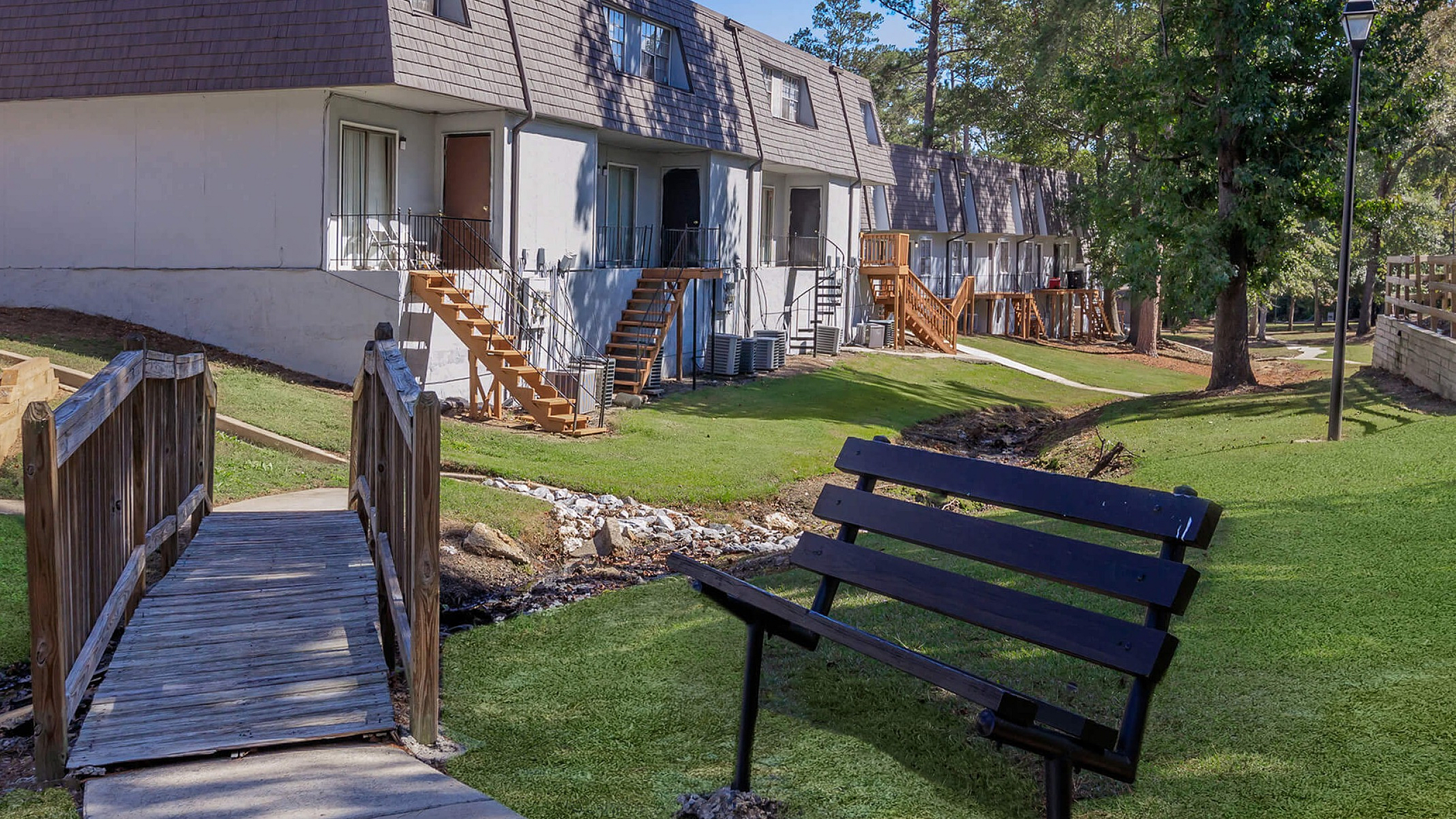 Quaint apartment complex with lush green lawns, benches, and a wooden bridge, surrounded by mature trees.