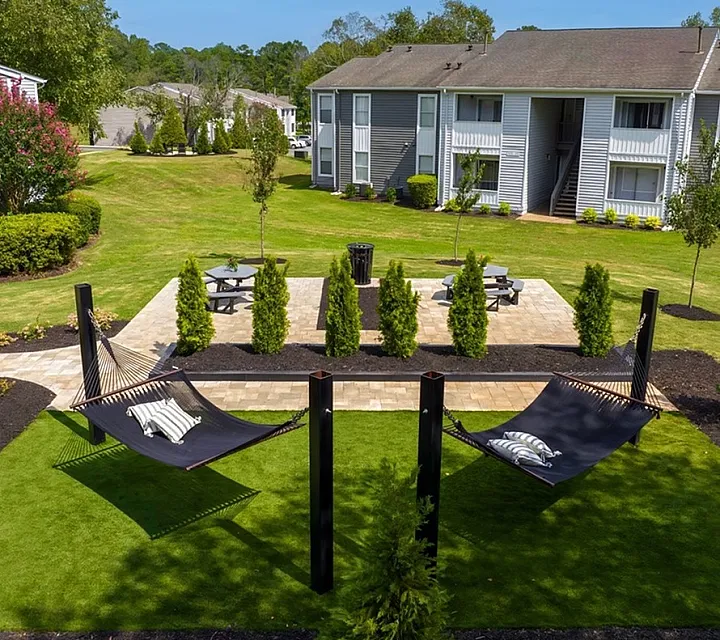 Lush outdoor area with hammocks, picnic tables, and cozy apartment buildings under a clear blue sky.