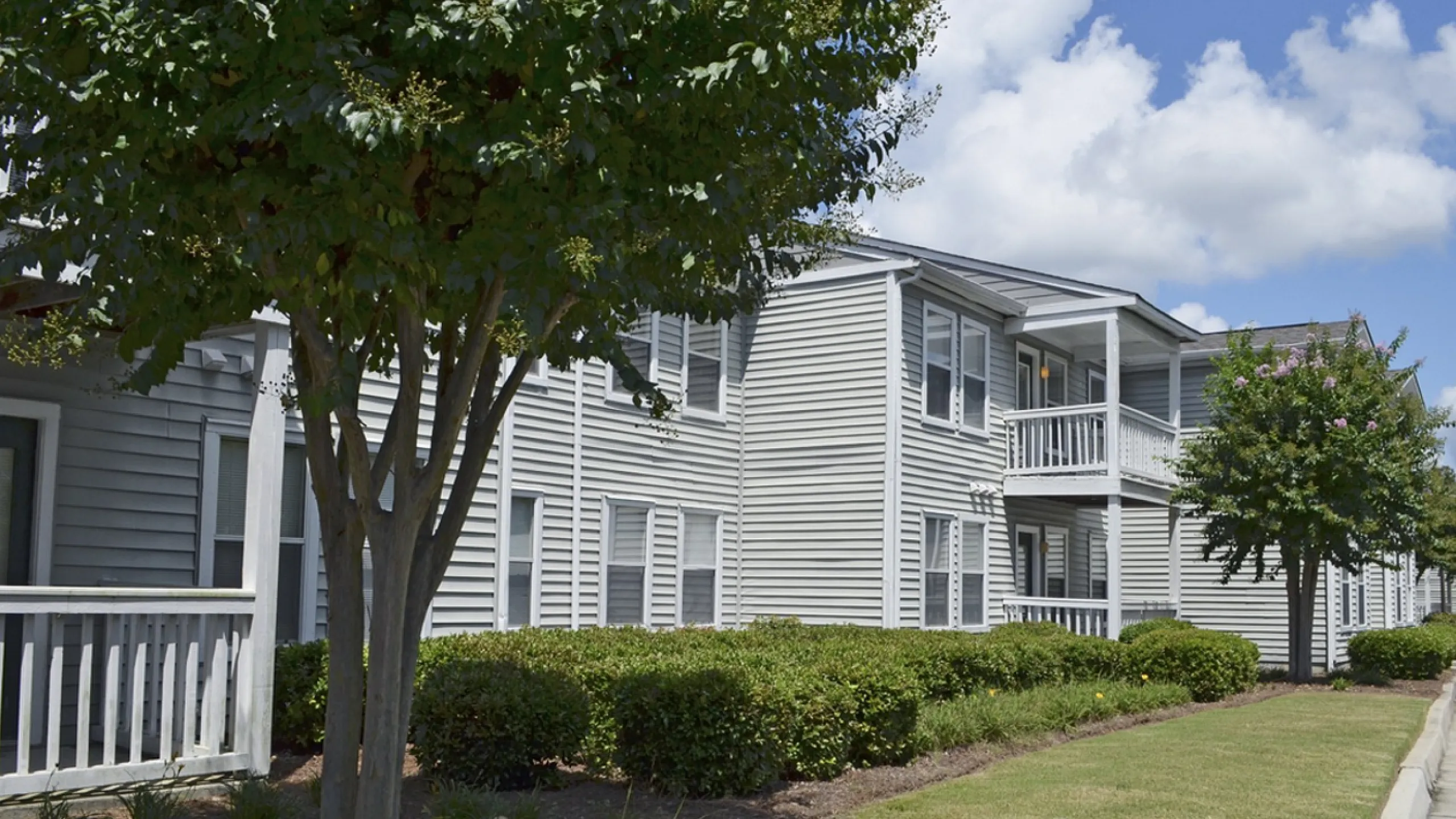 Charming apartment complex with lush greenery, private balconies, and a serene outdoor setting under a blue sky.