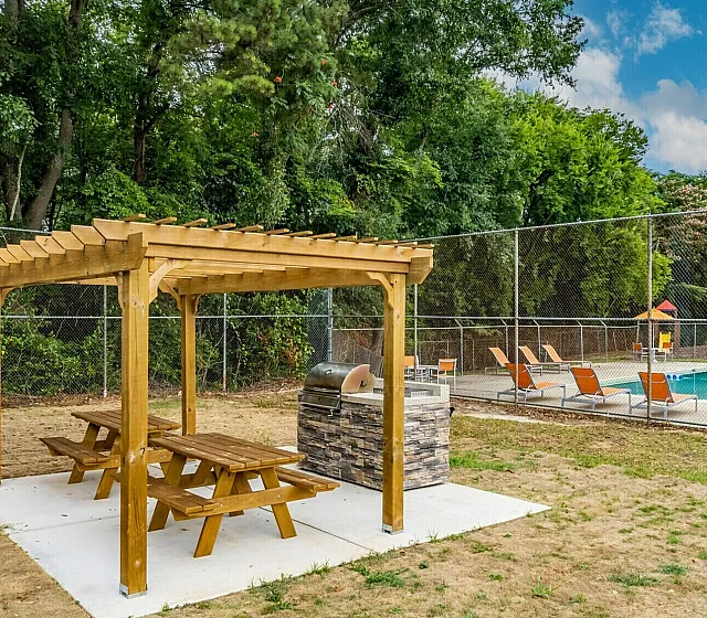 Inviting outdoor area with a wooden pergola and picnic bench beside a sparkling swimming pool, surrounded by lush greenery and comfortable lounge chairs.