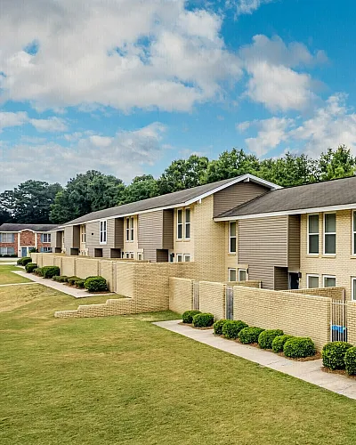 Charming row of townhouses with modern beige exteriors, private front patios, and lush green lawns under a bright blue sky.