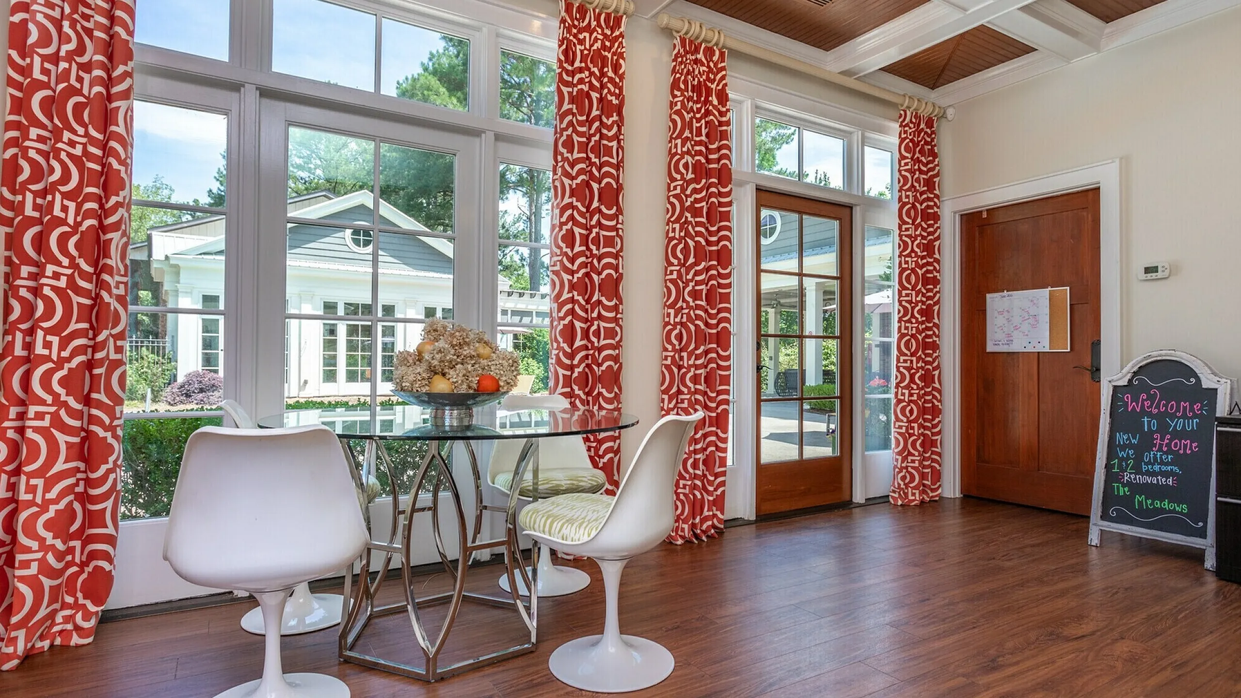 Bright, airy dining area with modern white chairs, elegant glass table, and vibrant red patterned curtains overlooking a lush garden.