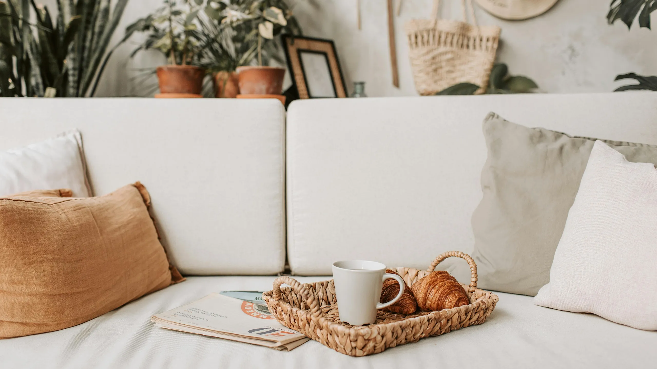 Cozy living space with a white sofa, decorative pillows, and a stylish wicker tray holding a cup and croissants. Lush green plants and chic wall decor create a serene ambiance.