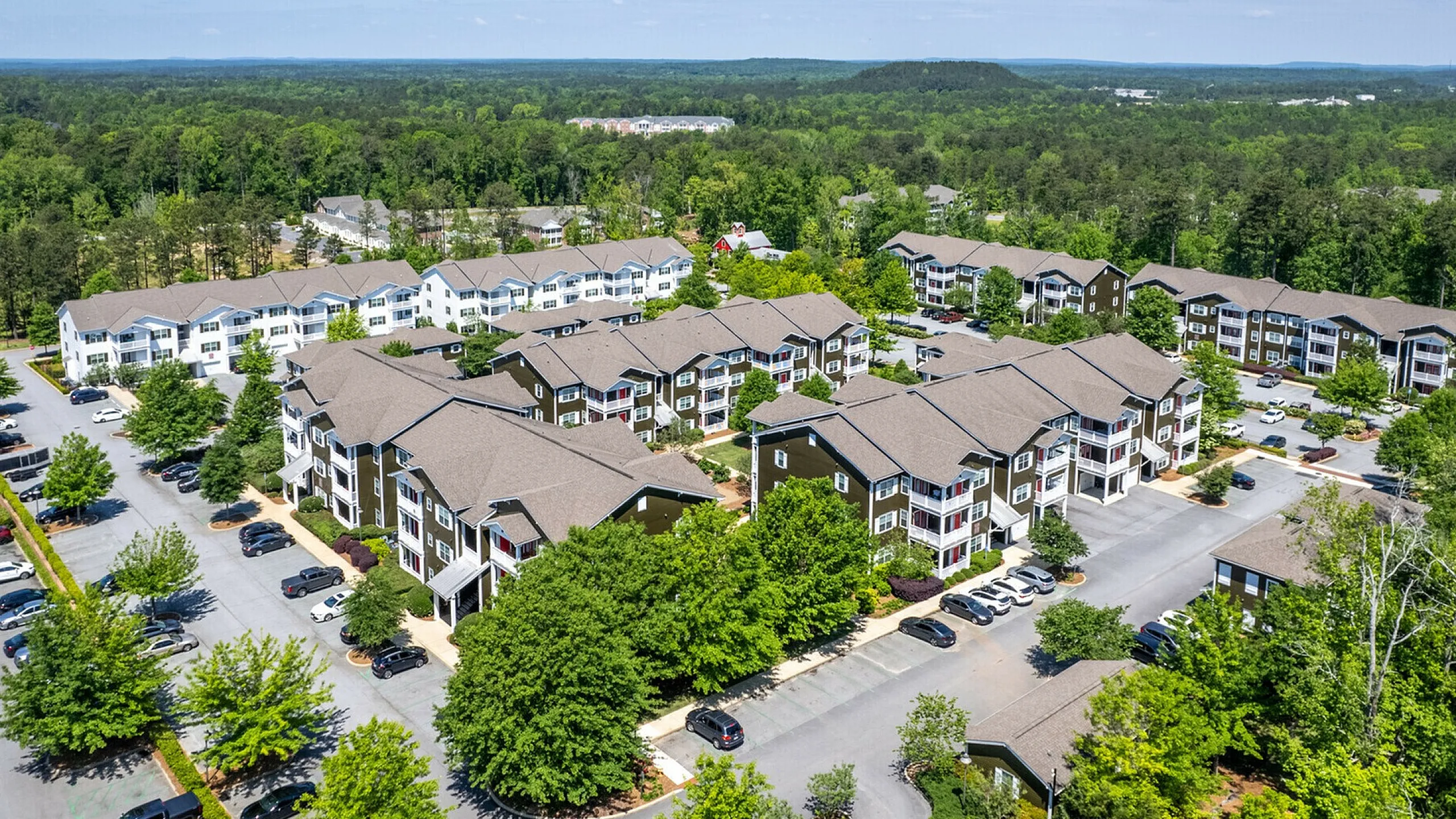 Aerial view of a beautiful apartment complex surrounded by lush greenery, showcasing modern architecture and ample parking facilities.