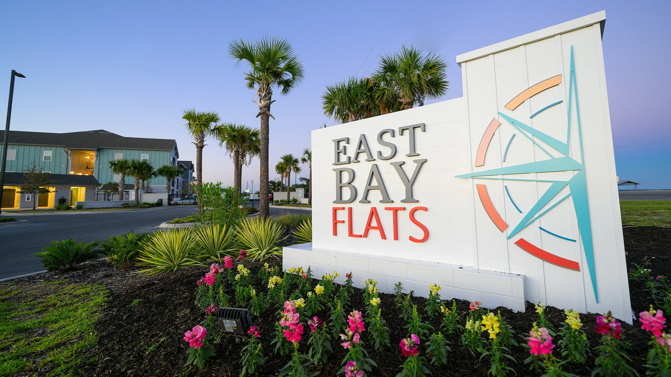 Inviting entrance to East Bay Flats with vibrant landscaping and modern design, perfectly set against a tranquil evening sky.