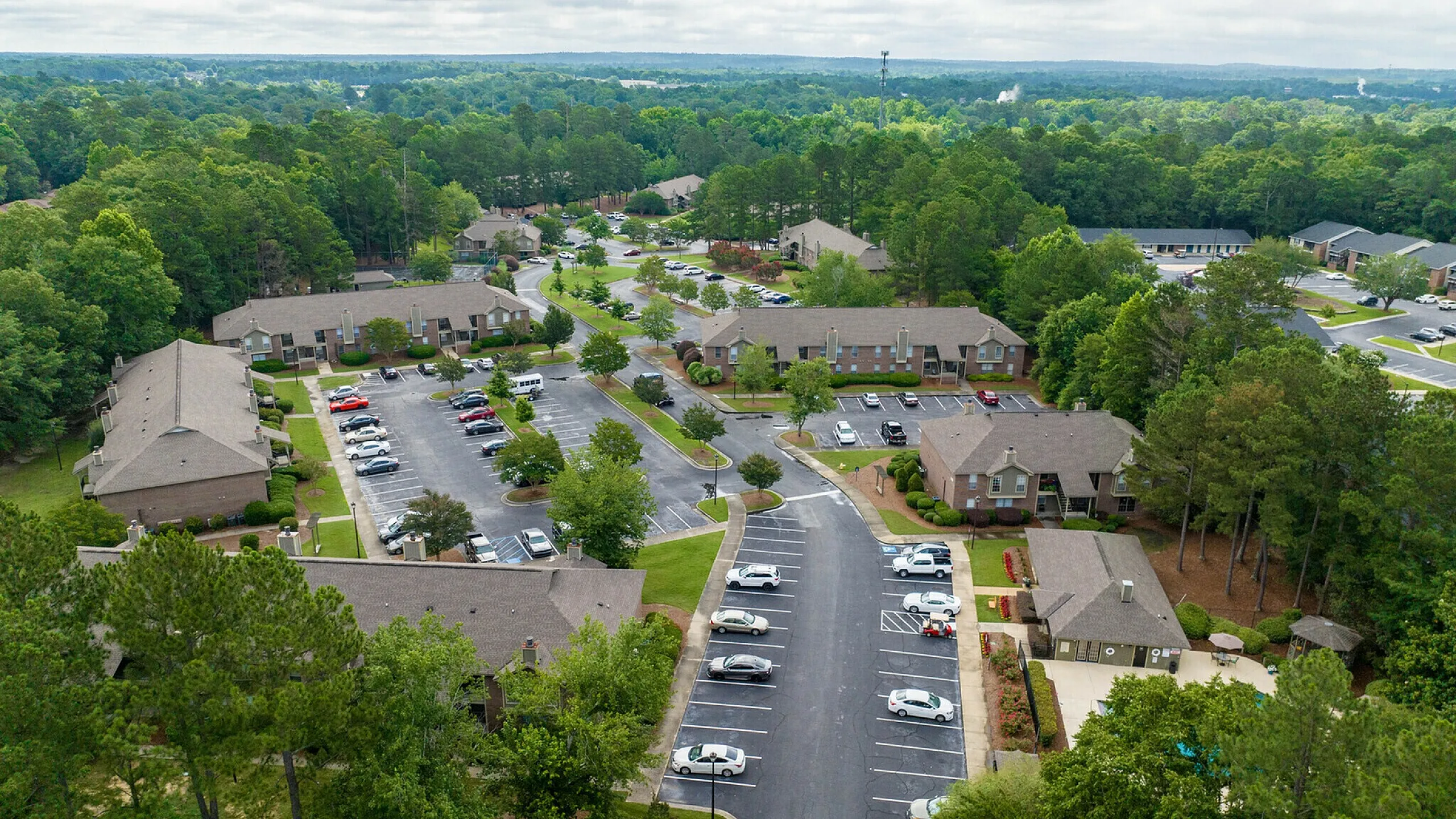 Aerial view of serene apartment complex nestled in lush greenery, featuring ample parking, charming pathways, and well-maintained landscaping.