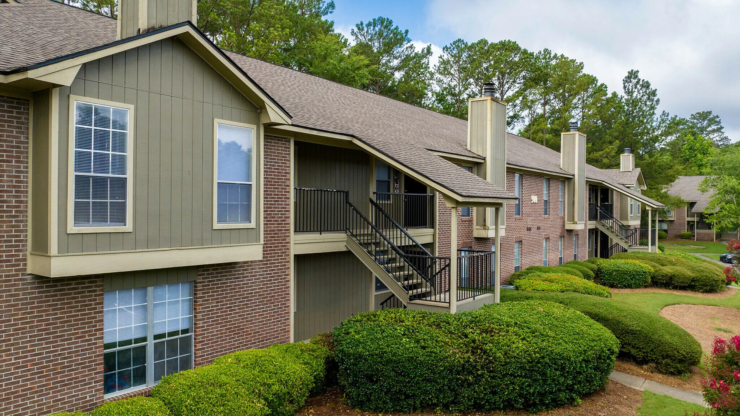 Charming apartment building with brick and wood accents, nestled among lush green landscaping and vibrant shrubbery, under a clear blue sky.