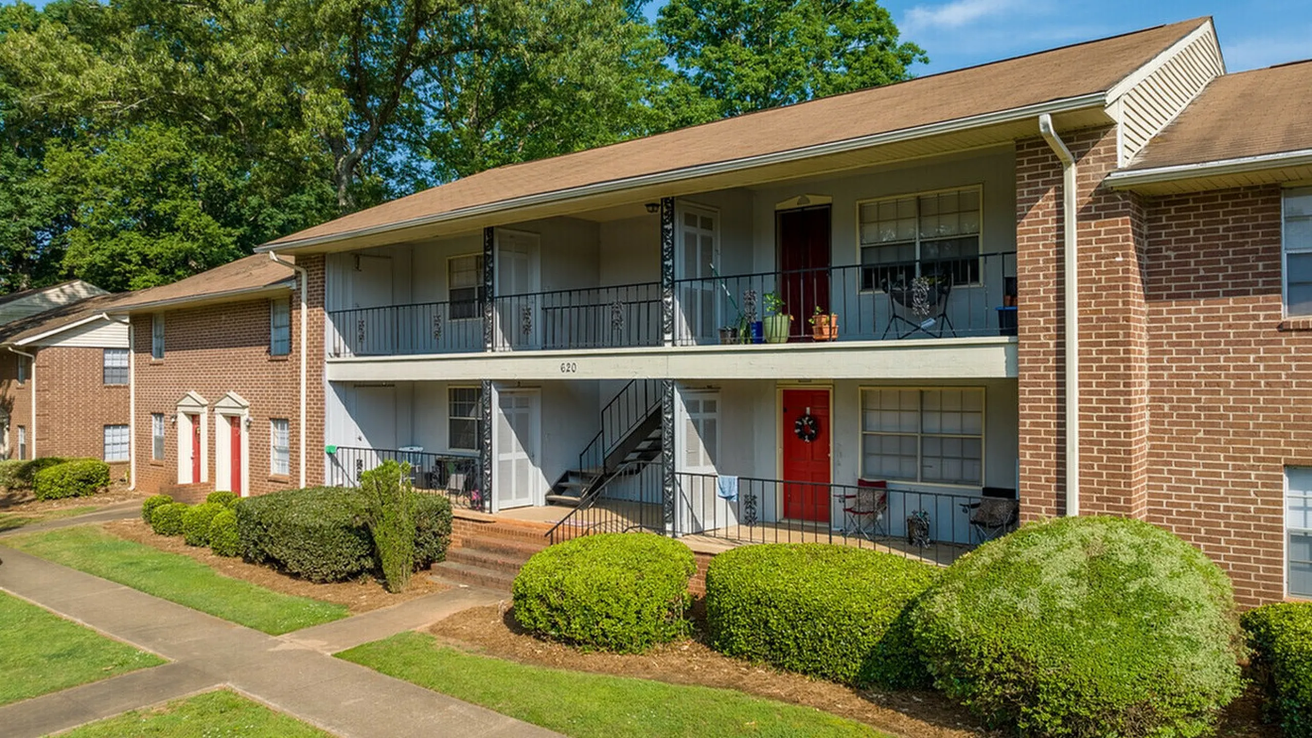 Charming two-story apartment building with spacious balconies and vibrant red doors, surrounded by lush greenery and inviting pathways.
