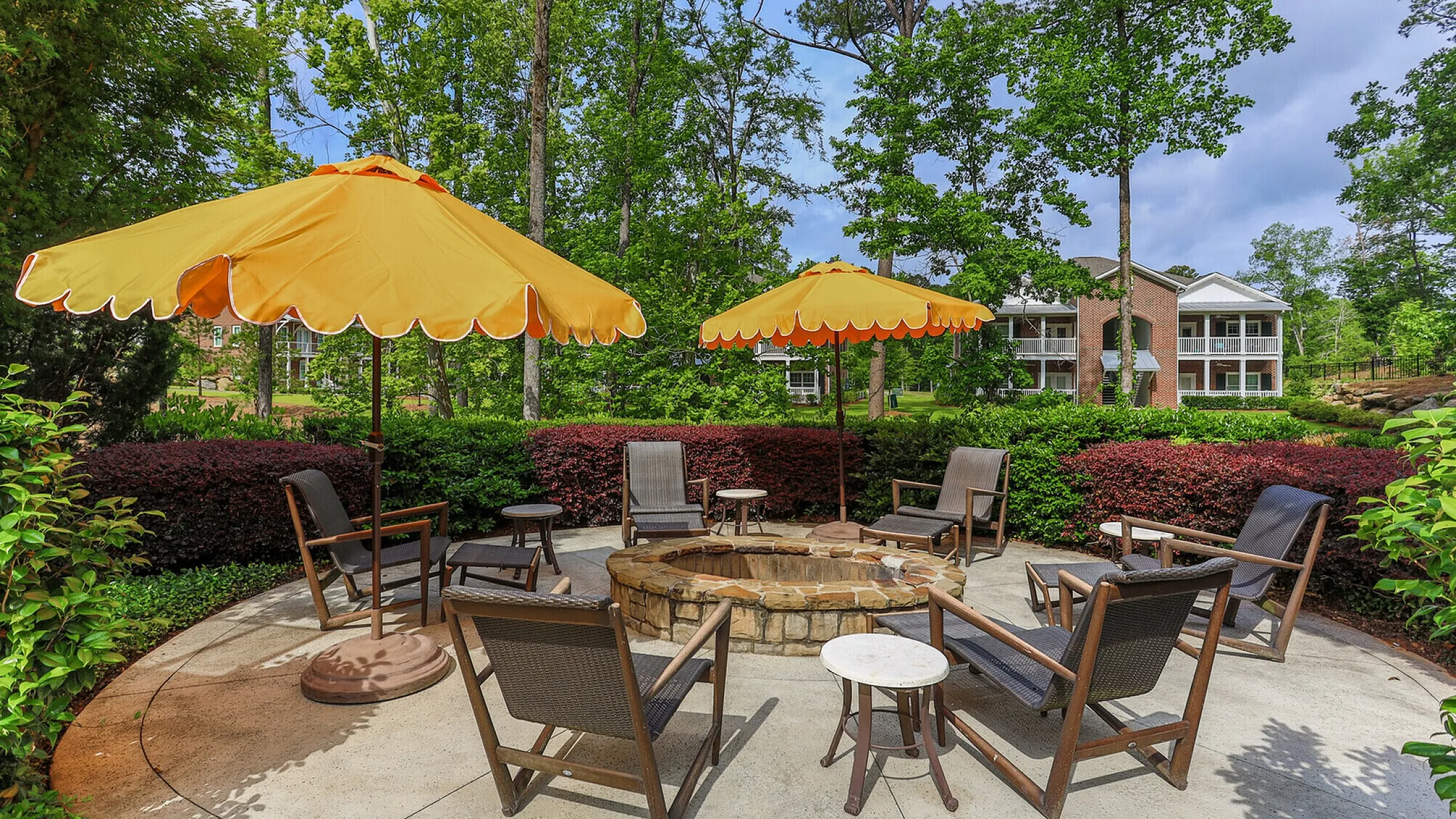 Charming outdoor patio with cozy fire pit, yellow umbrellas, and comfortable seating surrounded by lush greenery and scenic views of modern apartment buildings.