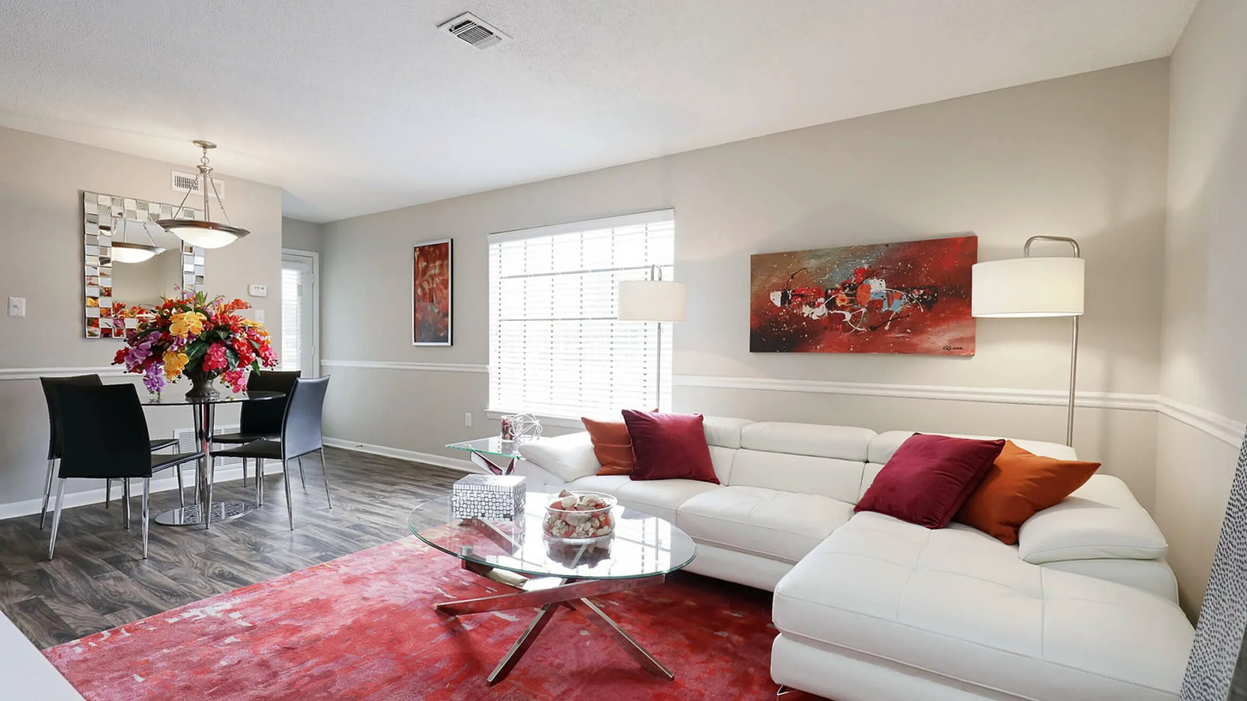 Chic living and dining area featuring a luxurious white sectional sofa, vibrant floral centerpiece, modern glass dining table, and bold red accents, perfect for stylish entertaining.