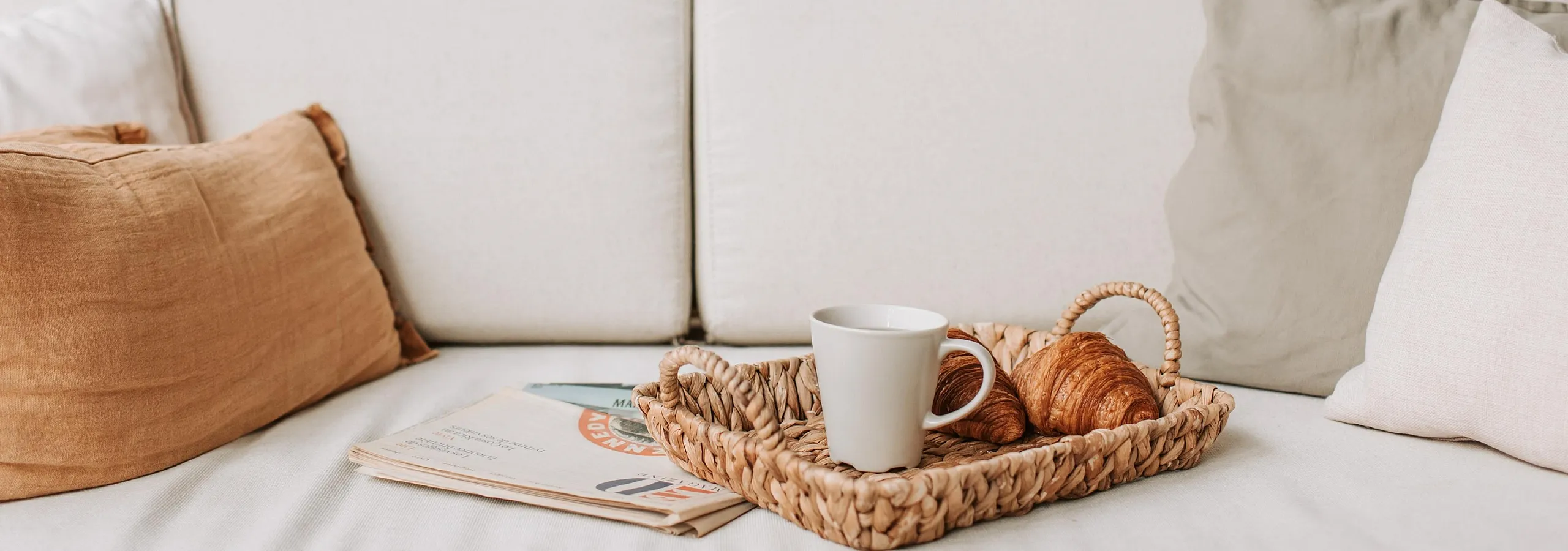 Cozy living room with plush cushions, surrounded by lush plants, featuring a stylish wicker tray with a cup and croissants for a relaxing ambiance.