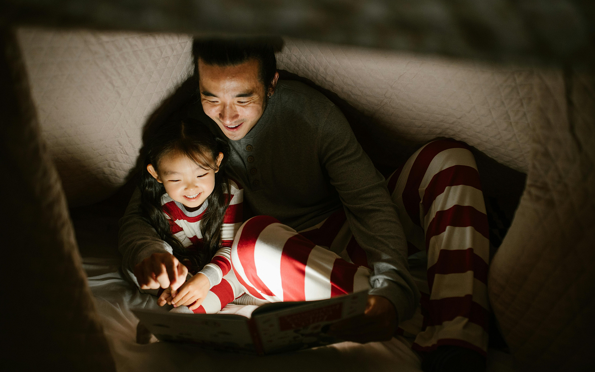 Cozy family moment inside a blanket fort, featuring a smiling parent and child in matching striped pajamas enjoying a bedtime story together.