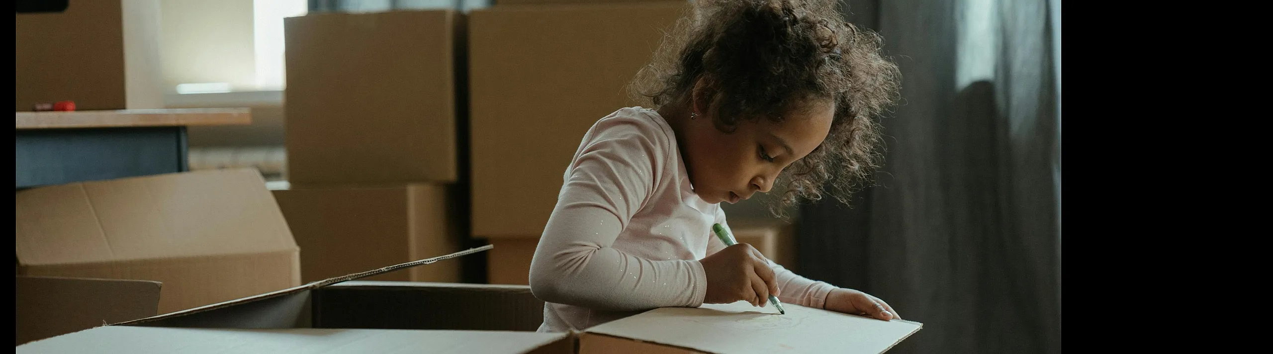 Charming scene of a child creatively coloring with vibrant hues inside a cozy cardboard box, surrounded by moving boxes in a sunlit room.