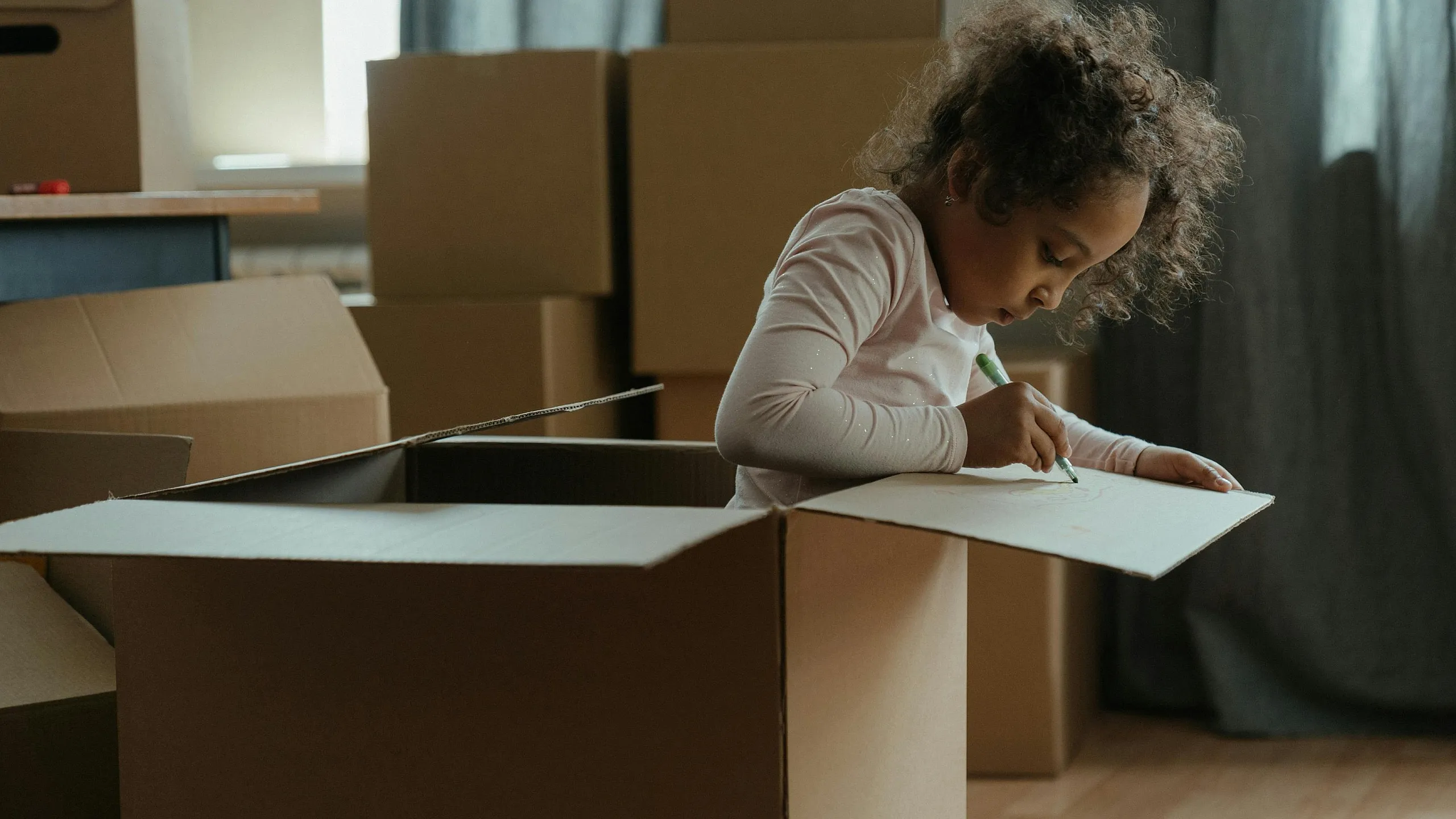 Cozy moving day scene with a child creatively drawing inside a cardboard box, surrounded by packing boxes and warm light.