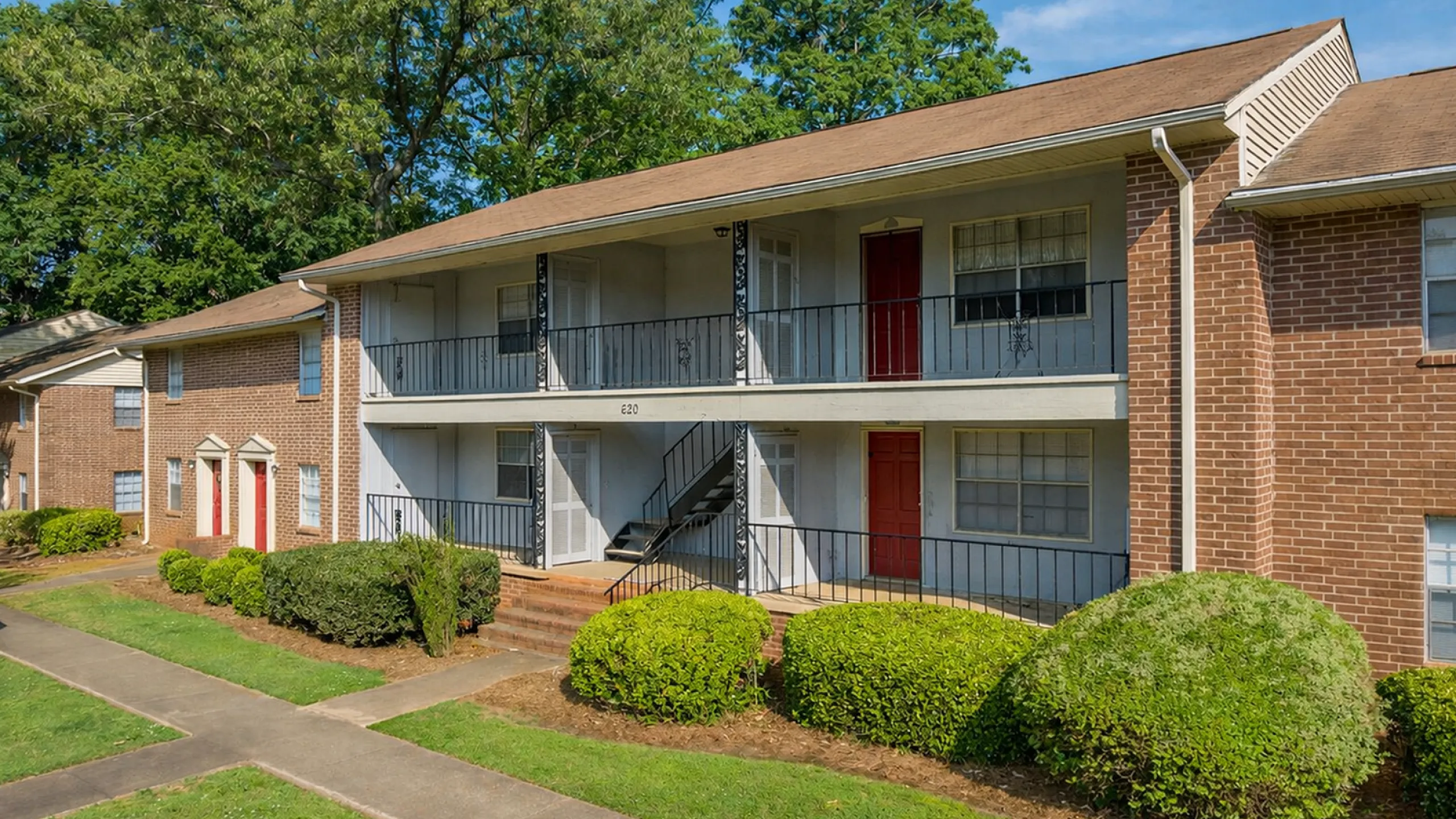 Charming brick apartment building with red doors, surrounded by lush greenery and shaded trees.