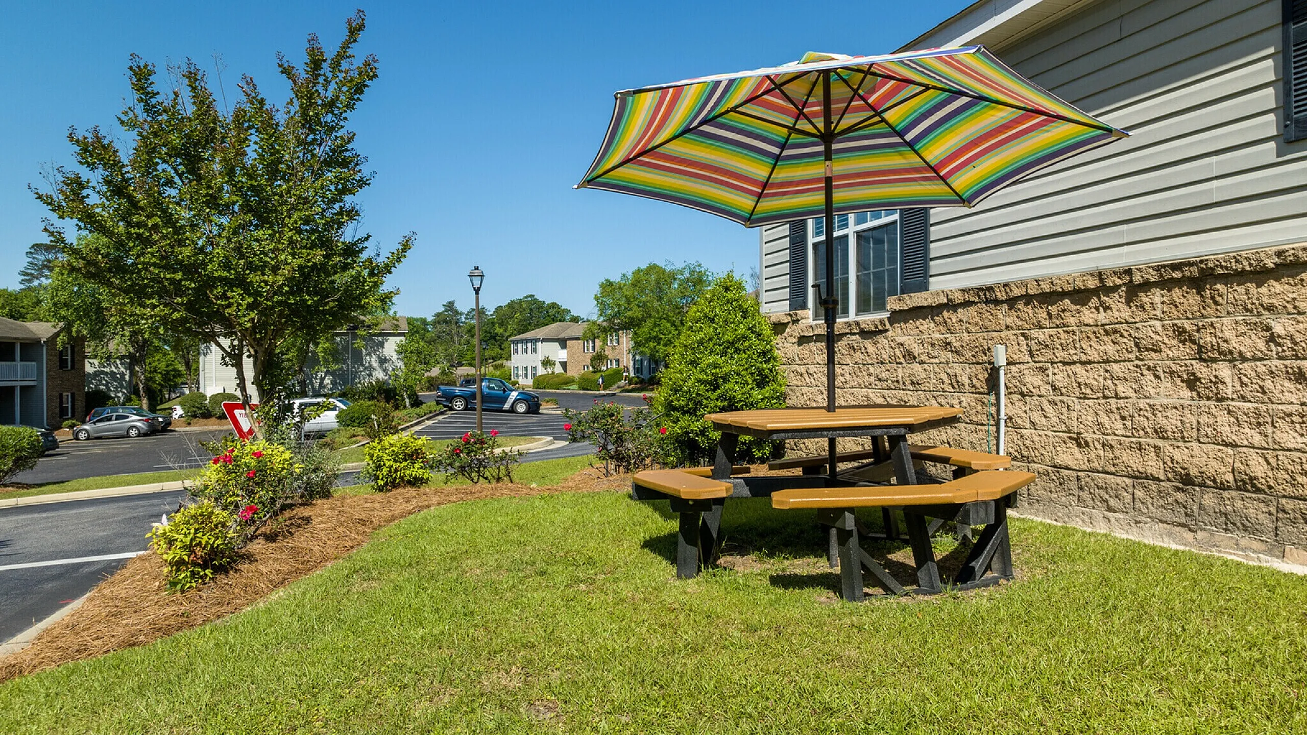 Inviting outdoor area with a vibrant umbrella and picnic table, surrounded by lush greenery and blooming flowers in a serene apartment community setting.