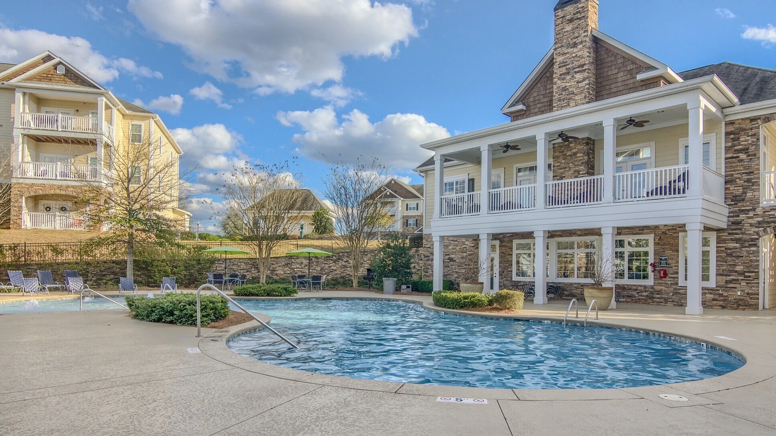 Inviting luxury apartment complex with a sparkling pool, modern stone architecture, and comfortable lounge chairs under a blue sky and fluffy clouds.
