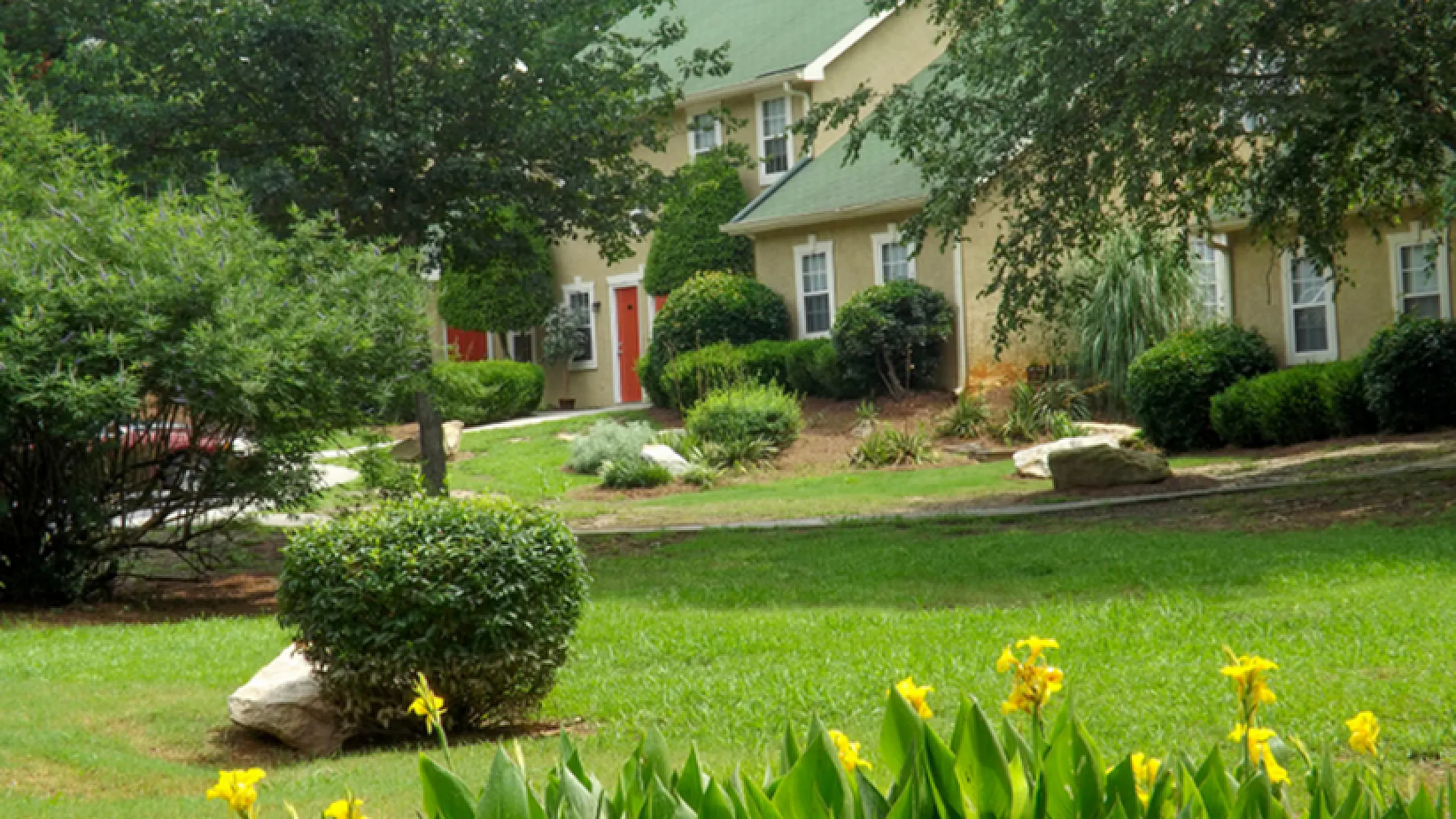 Charming suburban home surrounded by lush greenery and vibrant yellow flowers, featuring a cozy two-story design with a green roof and inviting red doors.