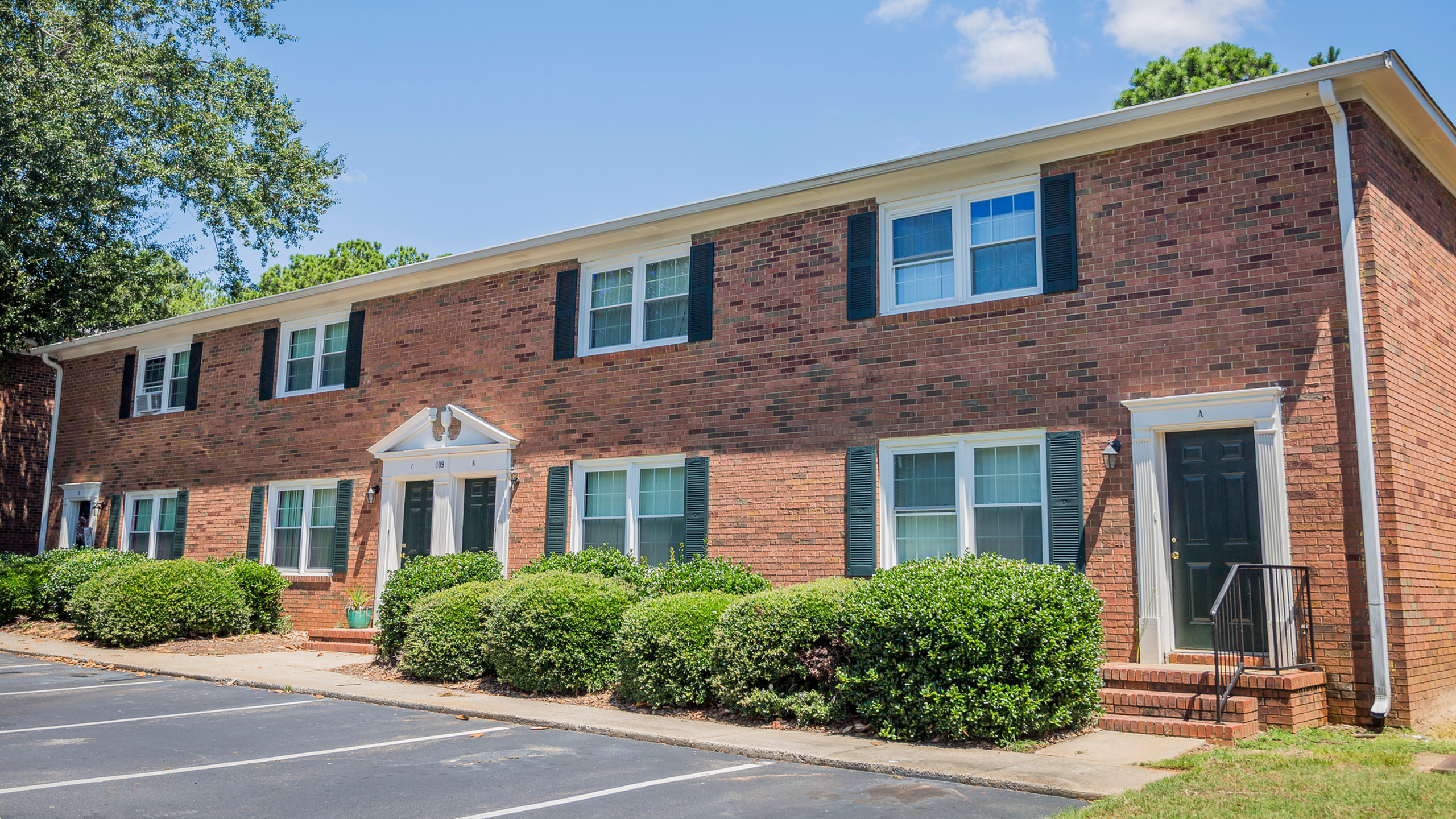 Charming brick apartment building with classic black shutters, surrounded by lush greenery and featuring convenient parking in front.