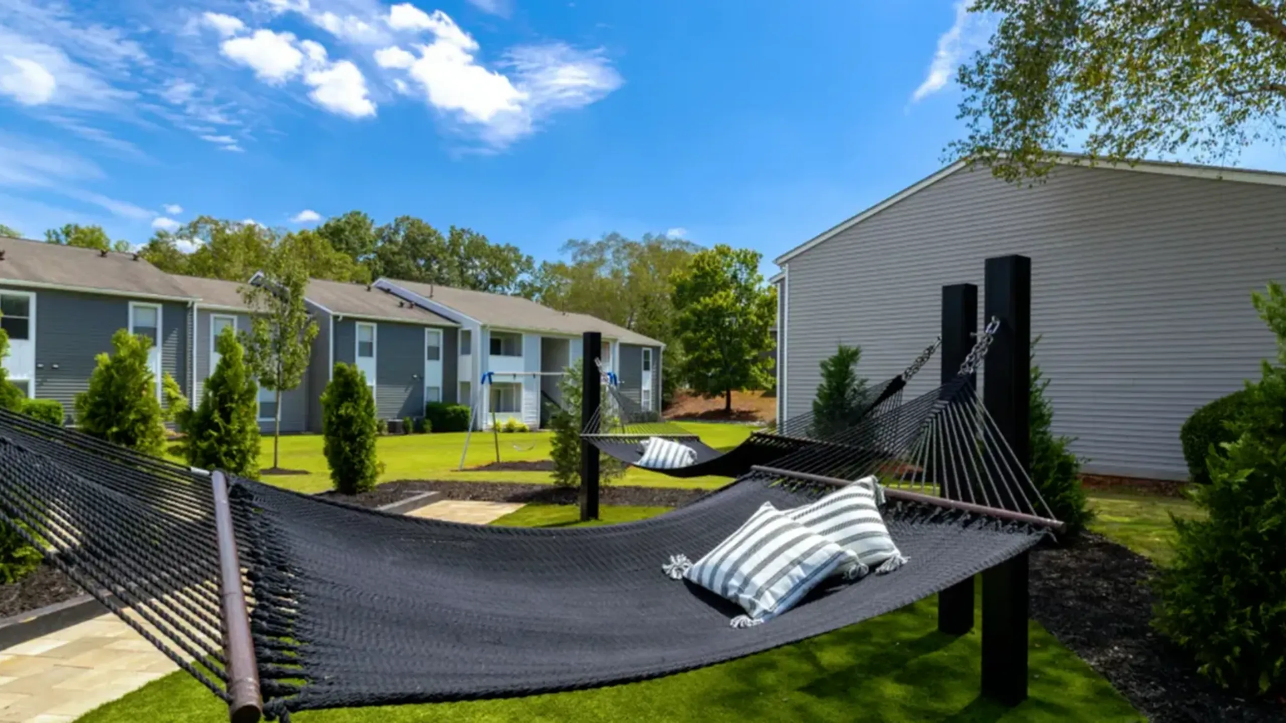 Serene outdoor area featuring cozy hammocks, lush green lawns, and modern apartments under a bright blue sky.