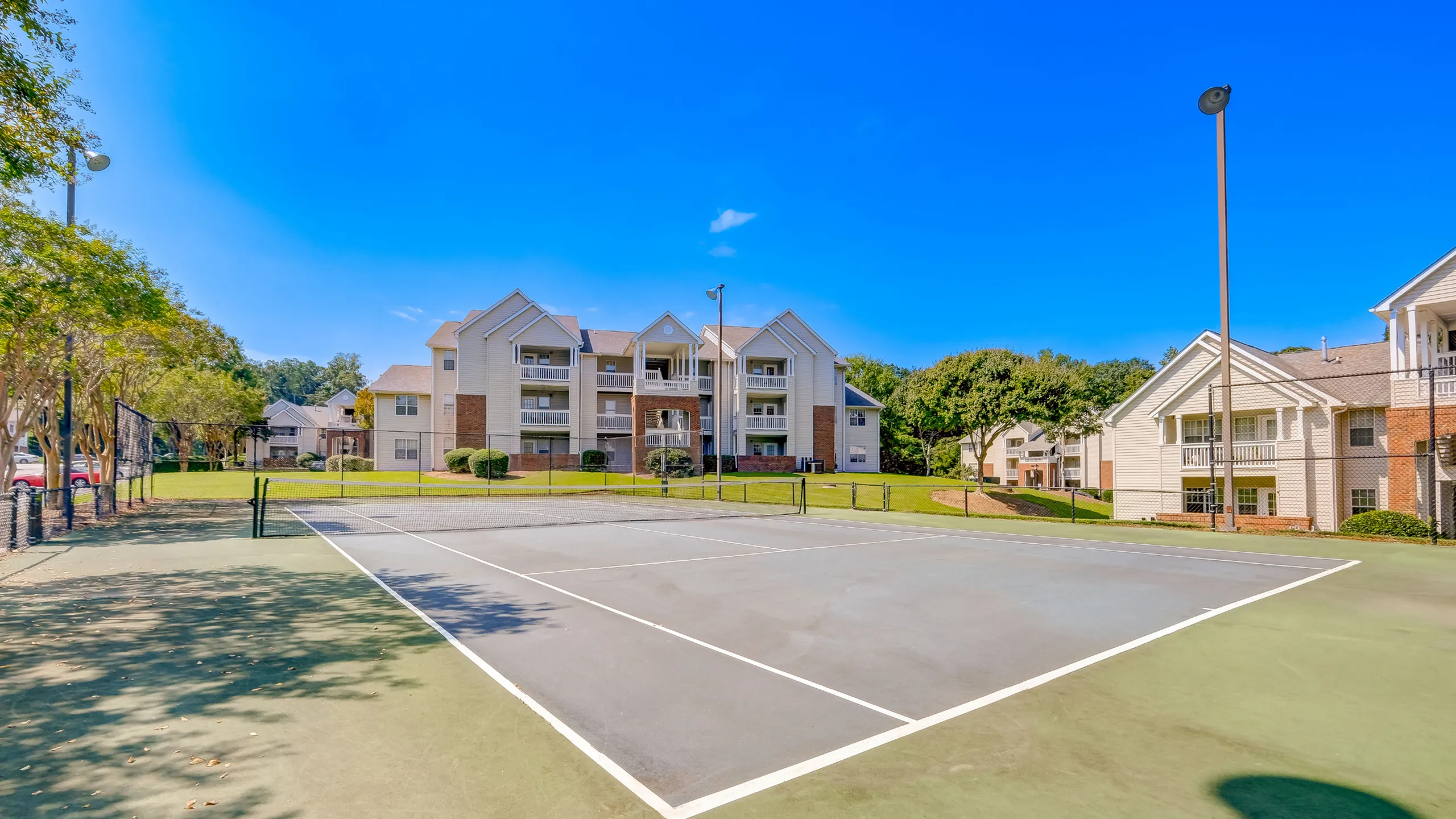 Charming apartment complex with elegant architecture, surrounded by lush greenery and a pristine tennis court under a clear blue sky.