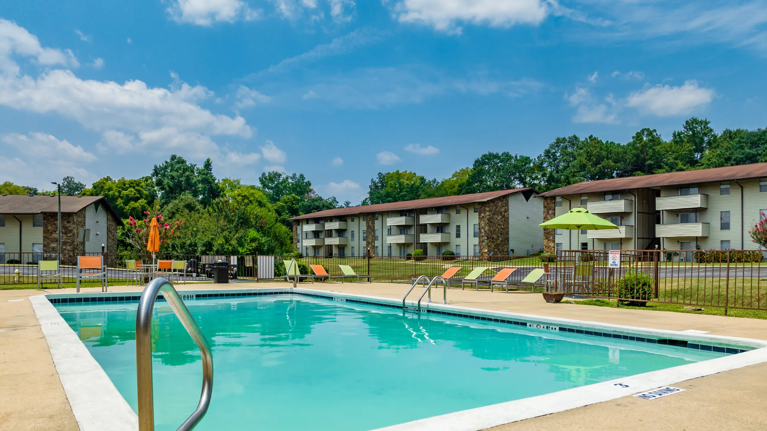 Inviting apartment complex featuring a sparkling swimming pool, surrounded by colorful lounge chairs and lush greenery under a bright blue sky.