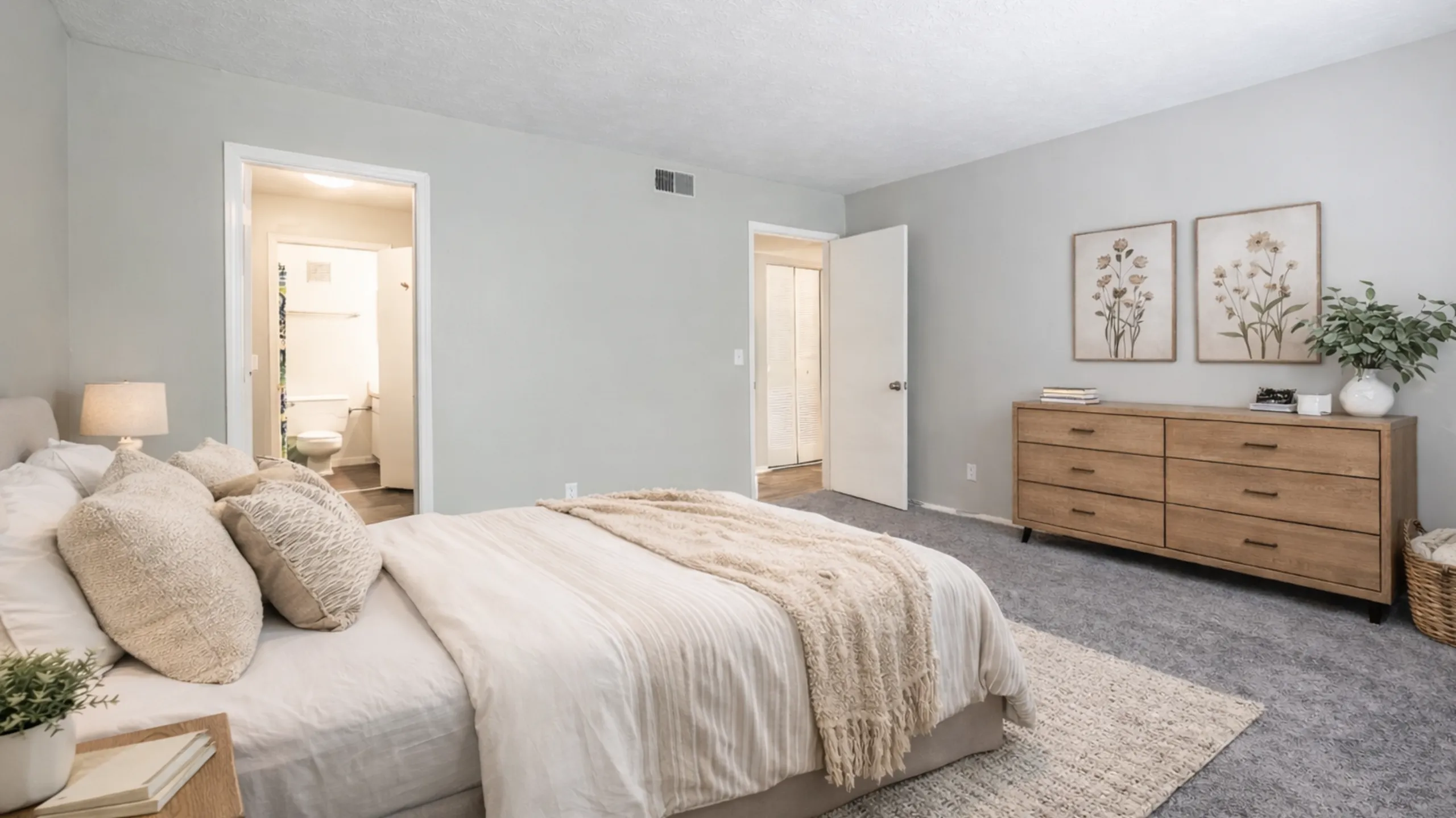 Elegant bedroom featuring a cozy queen bed with textured pillows, soft lighting, and a chic wooden dresser with floral art decor.