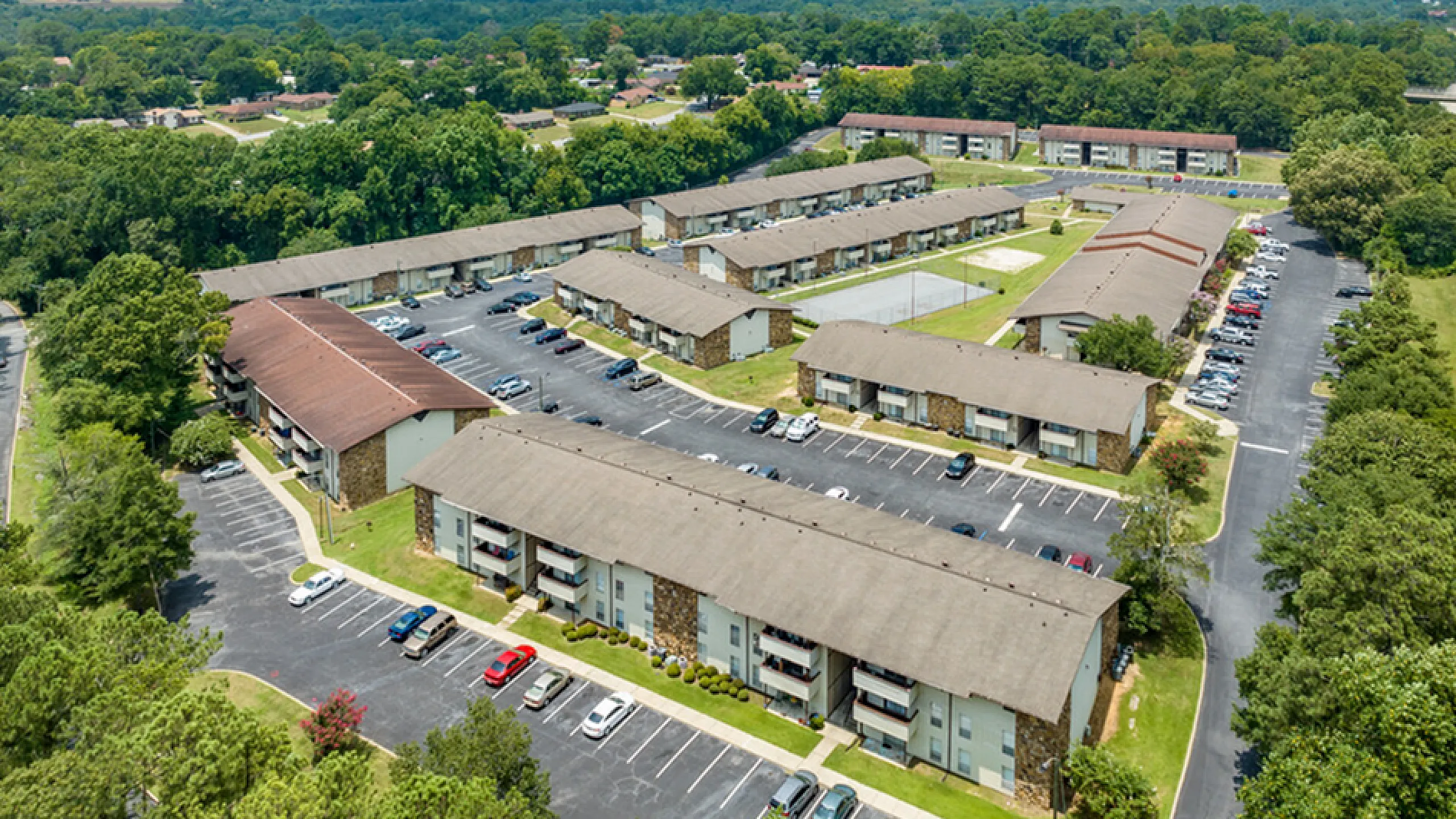 Aerial view of a vibrant apartment community surrounded by lush greenery, featuring modern buildings, ample parking, and recreational spaces.