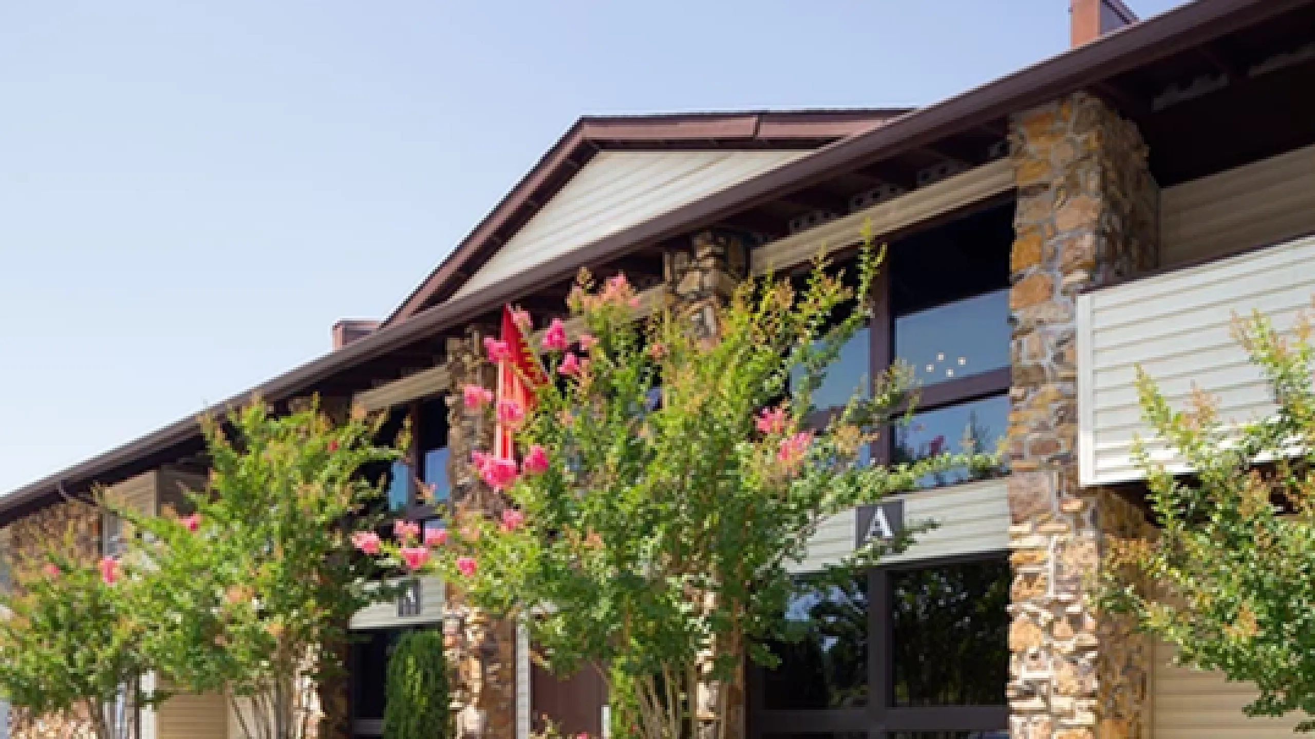 Charming apartment exterior with stone accents and lush landscaping, showcasing vibrant flowering trees under a clear blue sky.