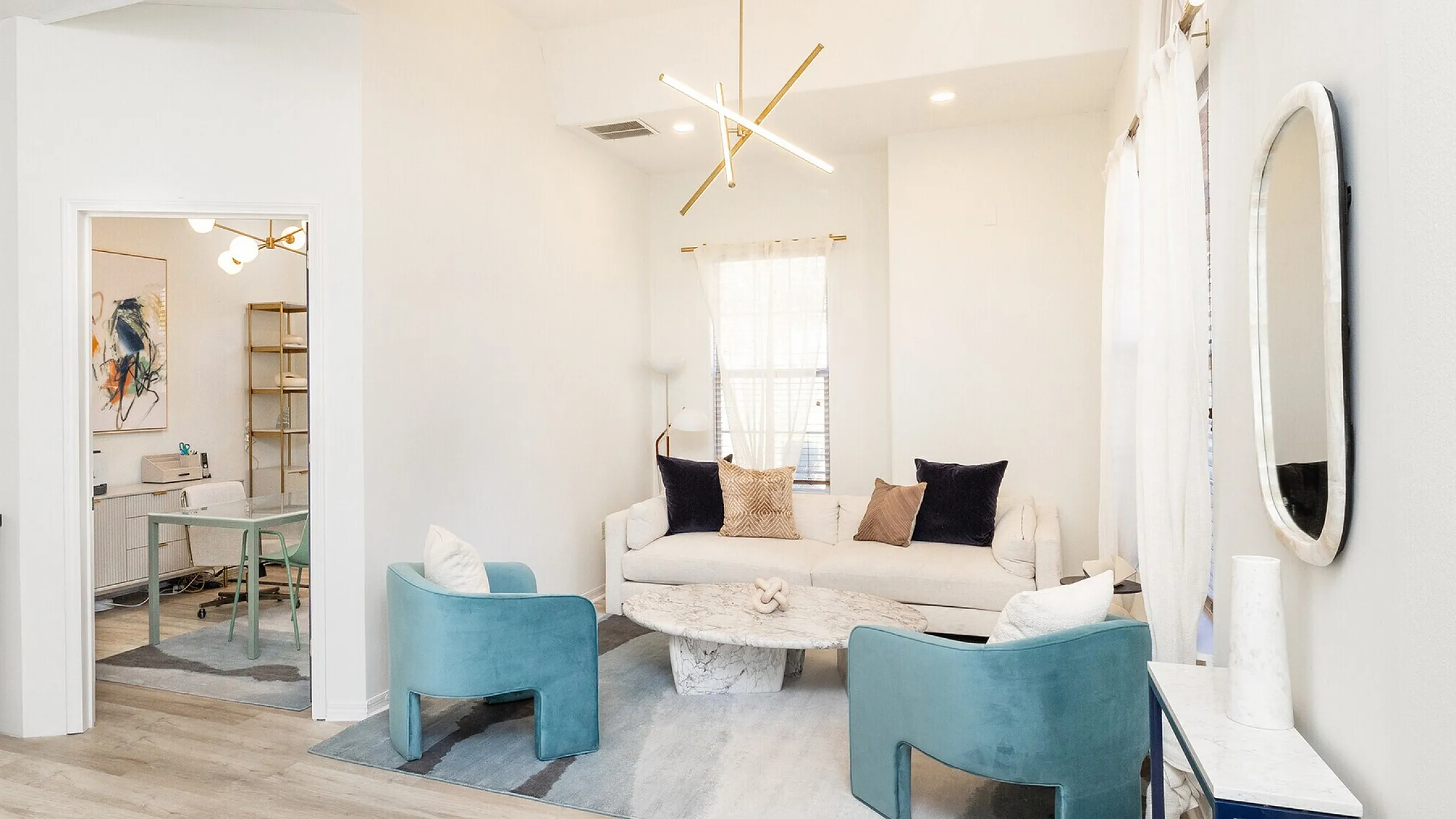 Chic living area with elegant sofa, marble coffee table, and plush blue chairs, bathed in natural light from large windows.