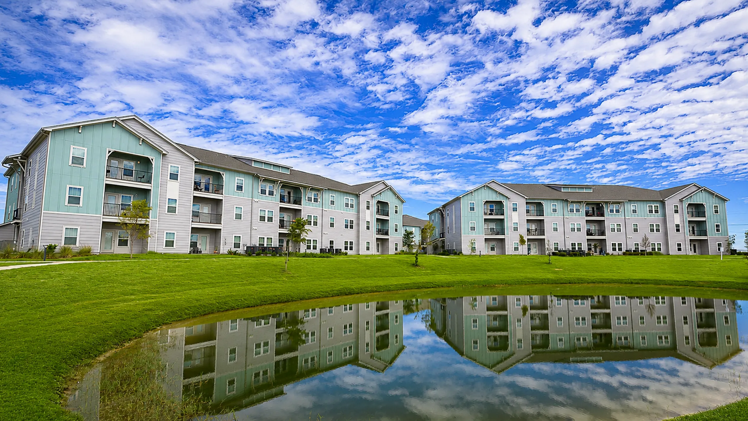 Charming apartment complex featuring modern architecture with lush green lawns and a tranquil pond reflecting a clear blue sky.