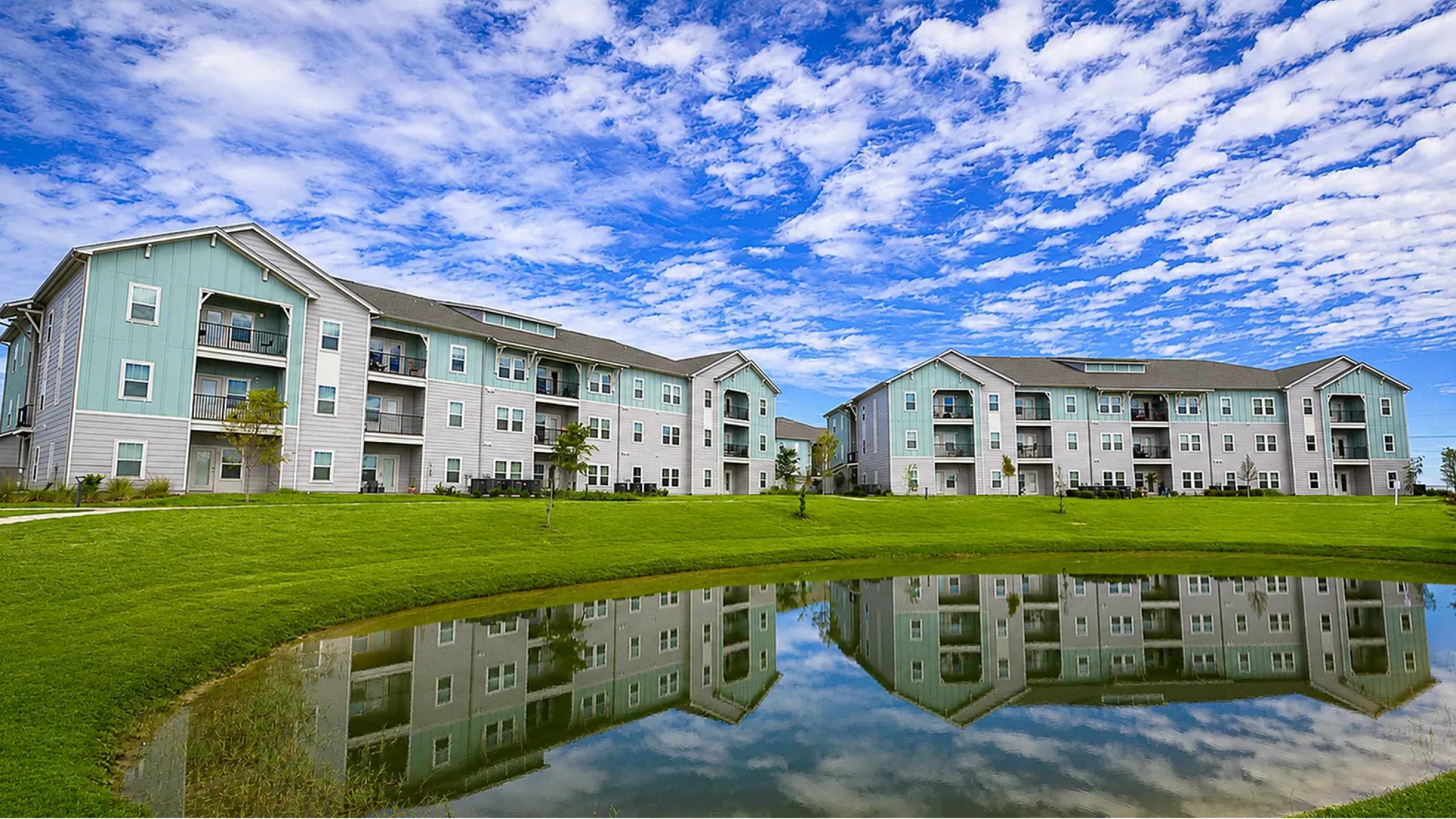 Charming apartment complex featuring modern architecture with serene pond reflections and expansive green spaces under a vibrant blue sky.