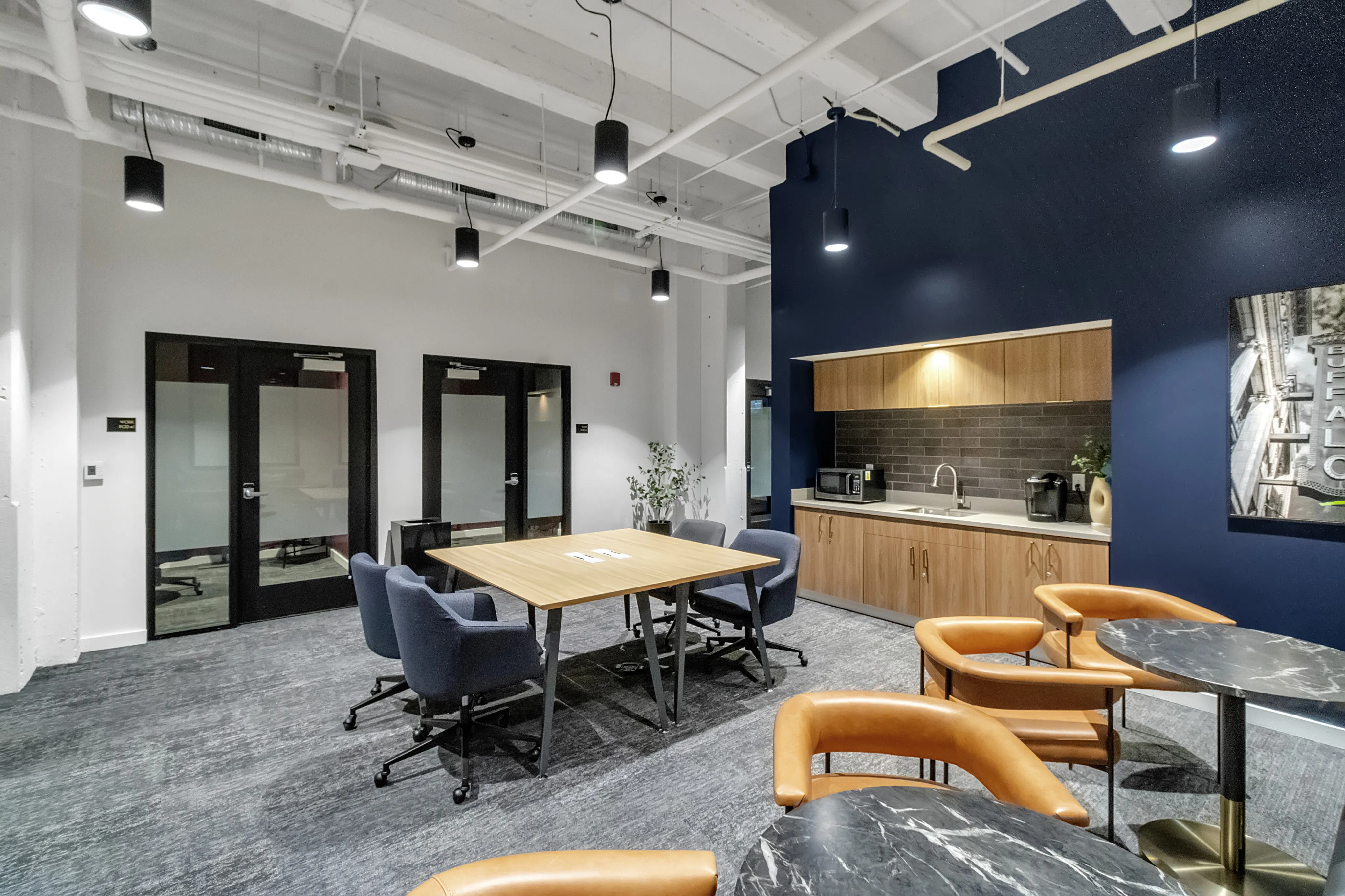 Modern and inviting office space featuring a stylish meeting area with sleek wooden table, comfortable blue chairs, and a chic kitchenette with a bold blue accent wall.